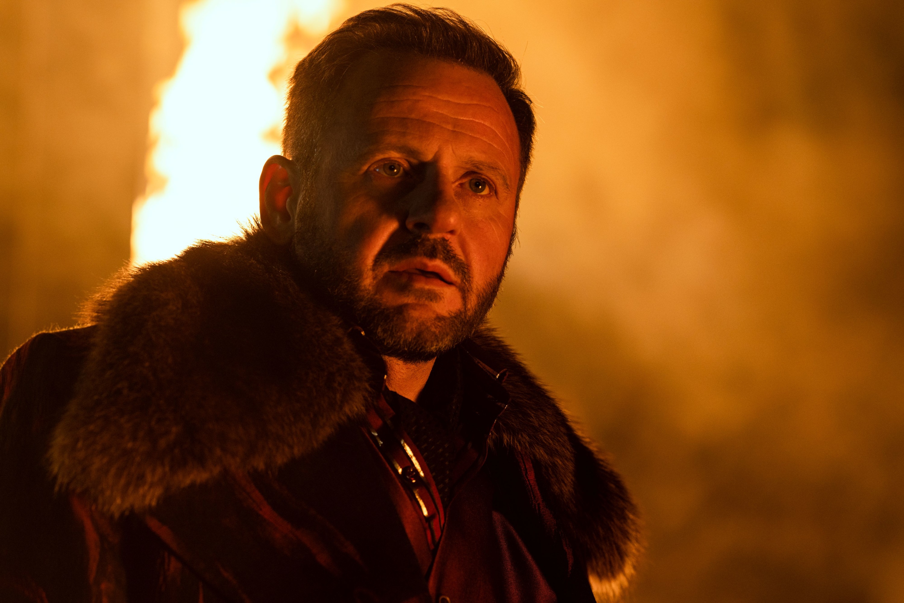 Man in fur collar coat in front of fire HD Wallpaper