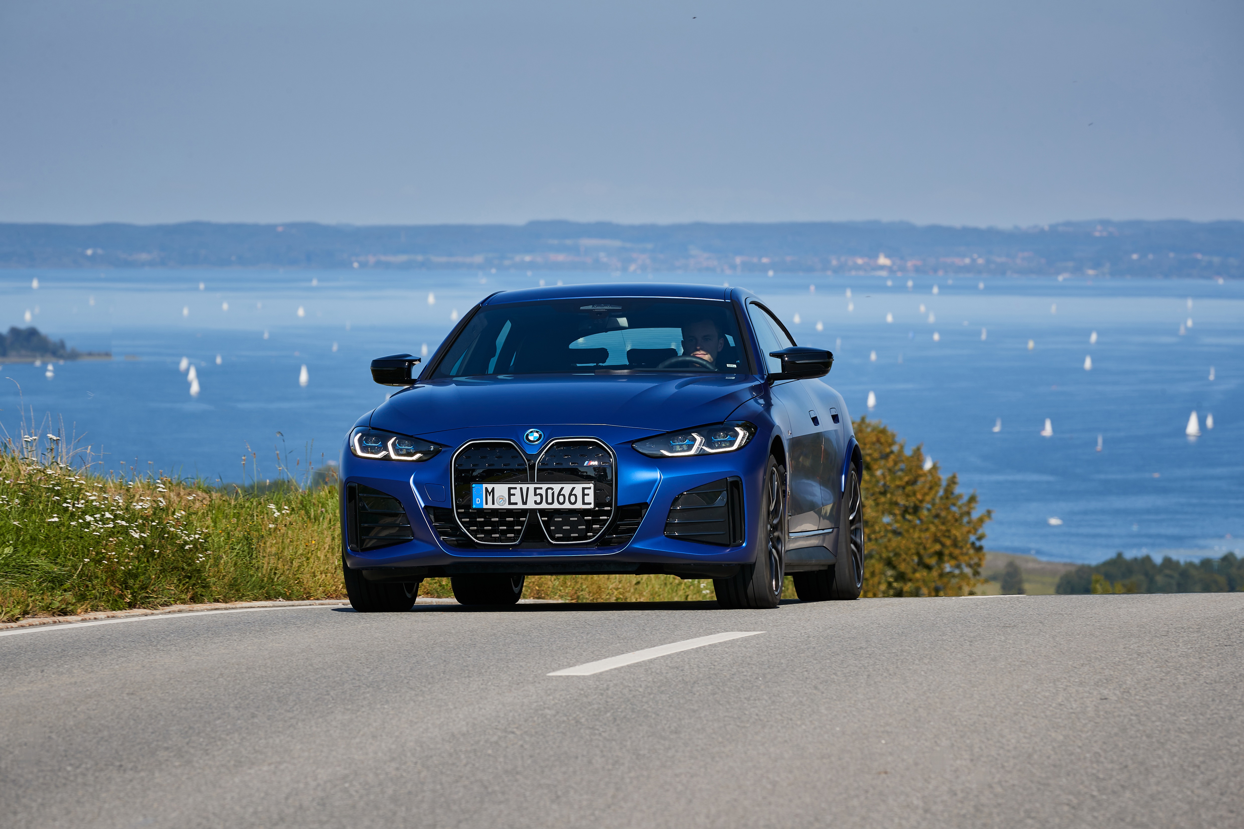 Blue BMW i4 M50 driving on a road with a lake in the background HD Wallpaper
