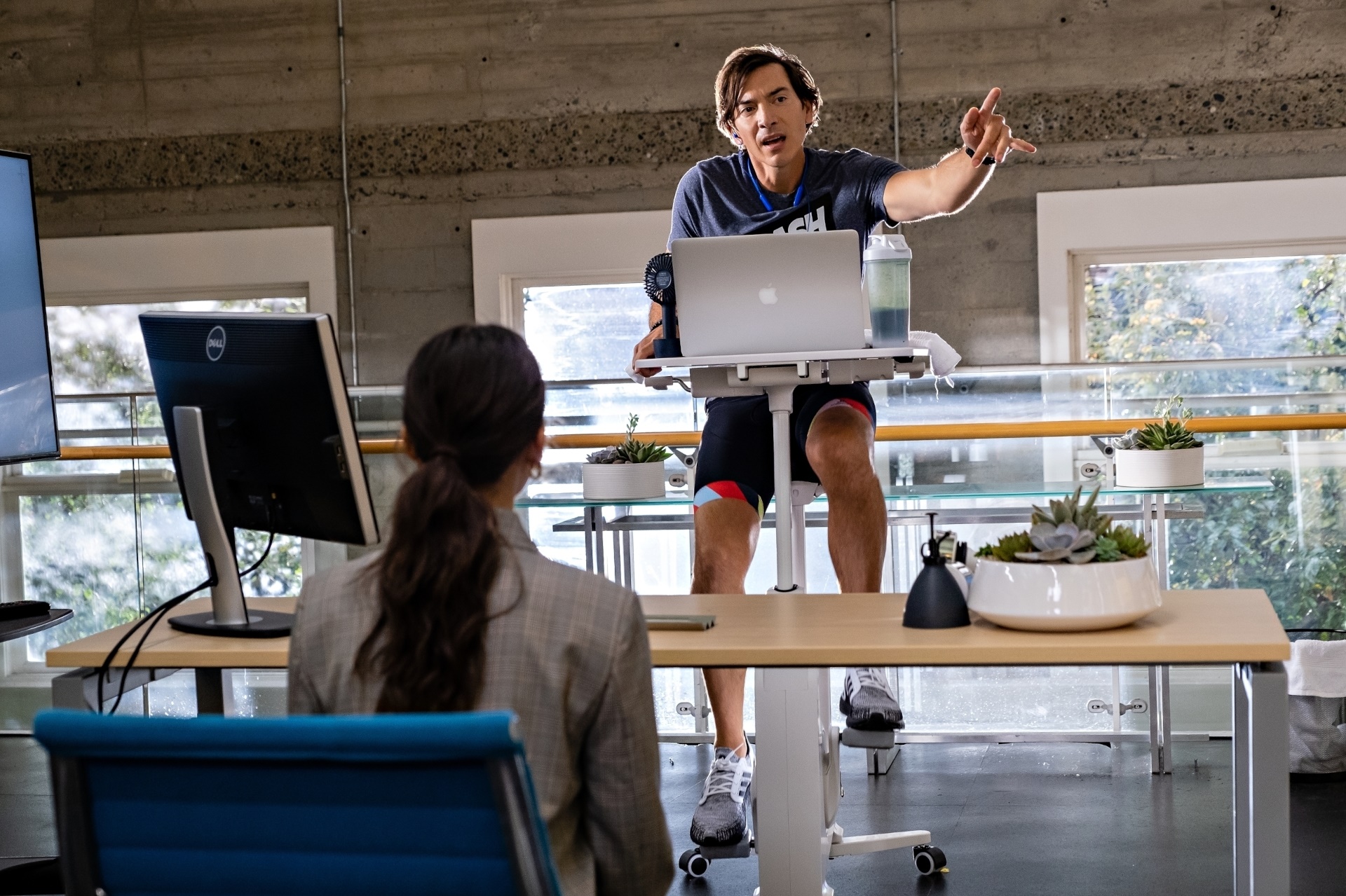 Man on stationary bike teaching woman at desk HD Wallpaper