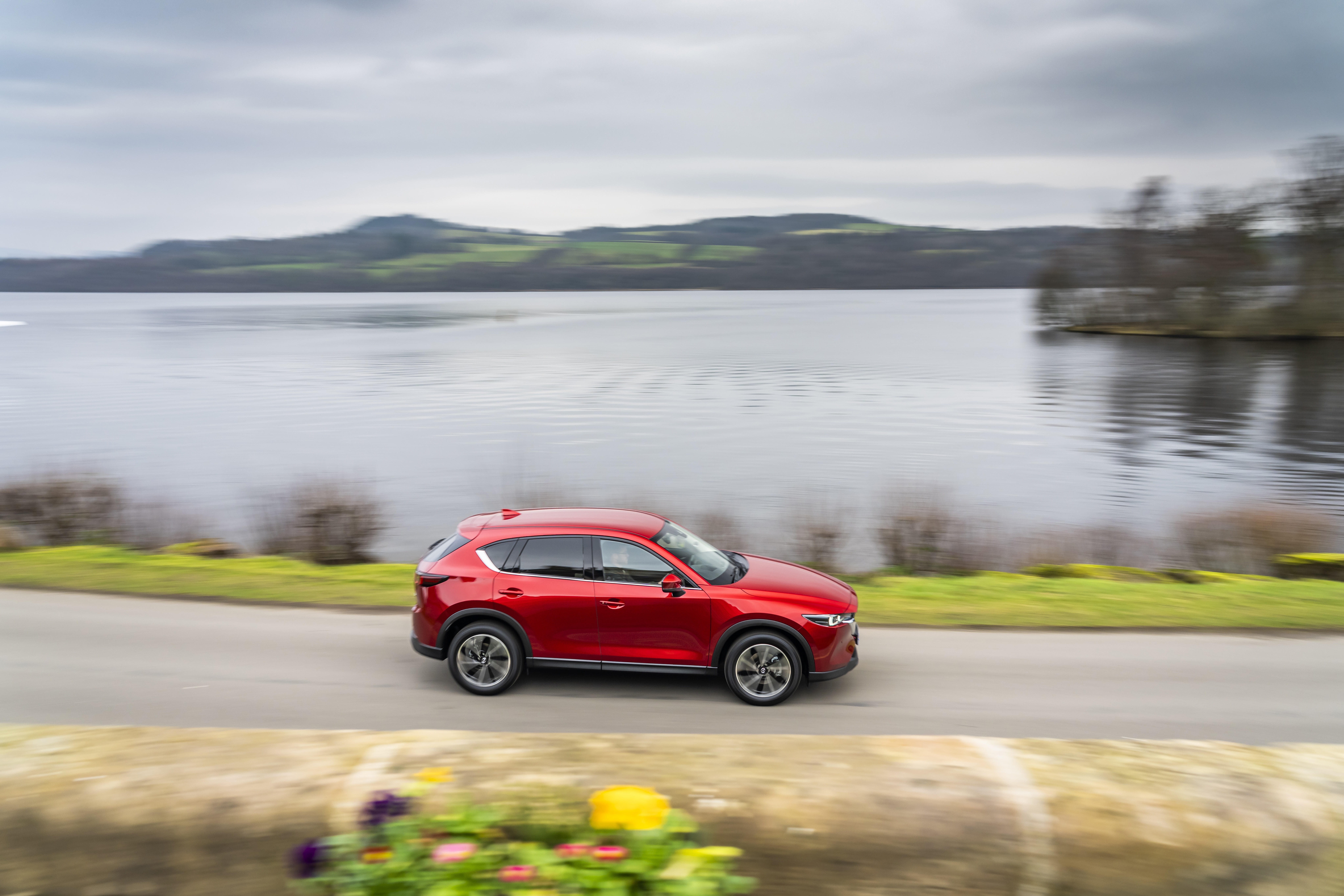 Red Mazda CX-5 driving on a road by a lake HD Wallpaper