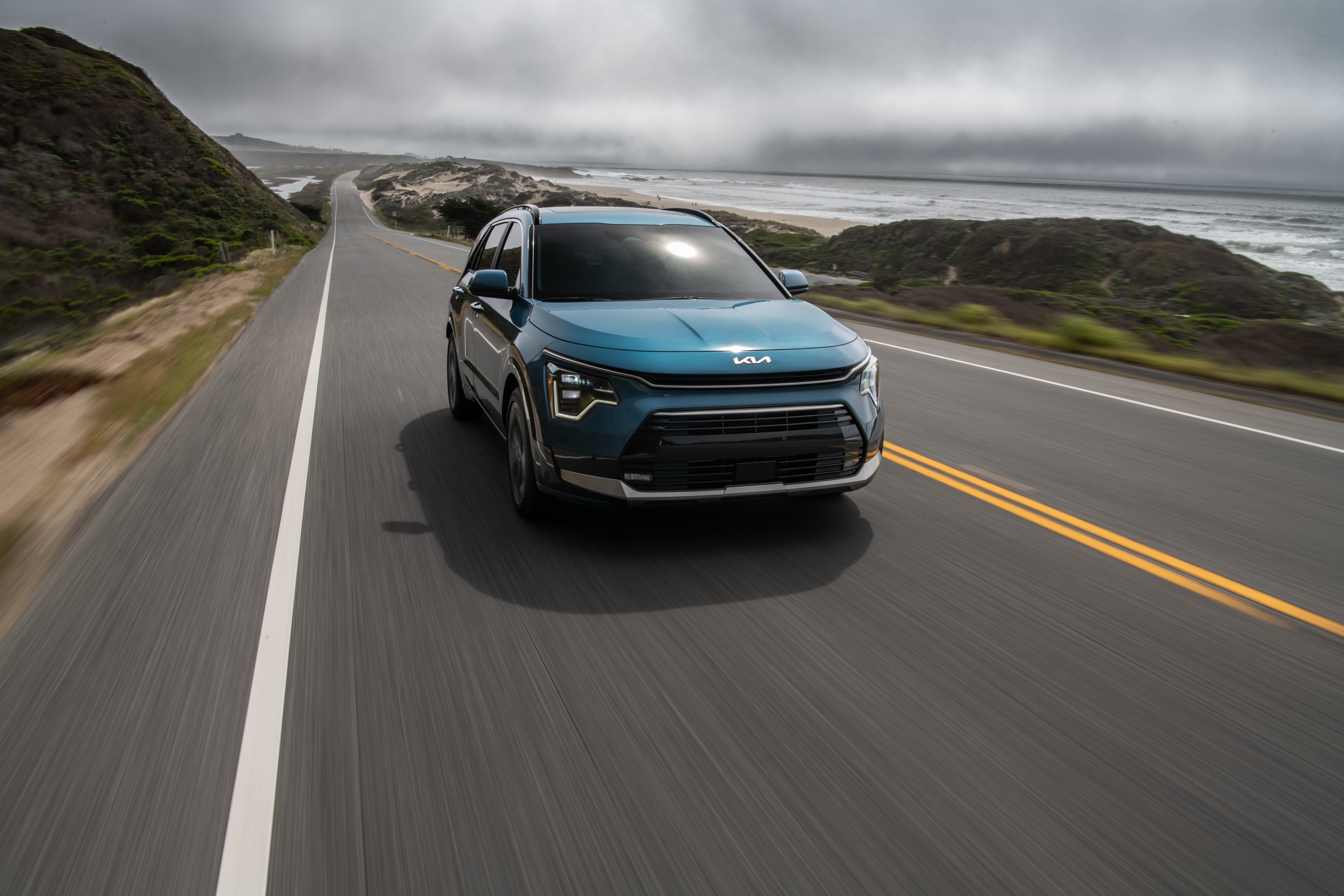 Blue Kia Niro driving on coastal highway HD Wallpaper