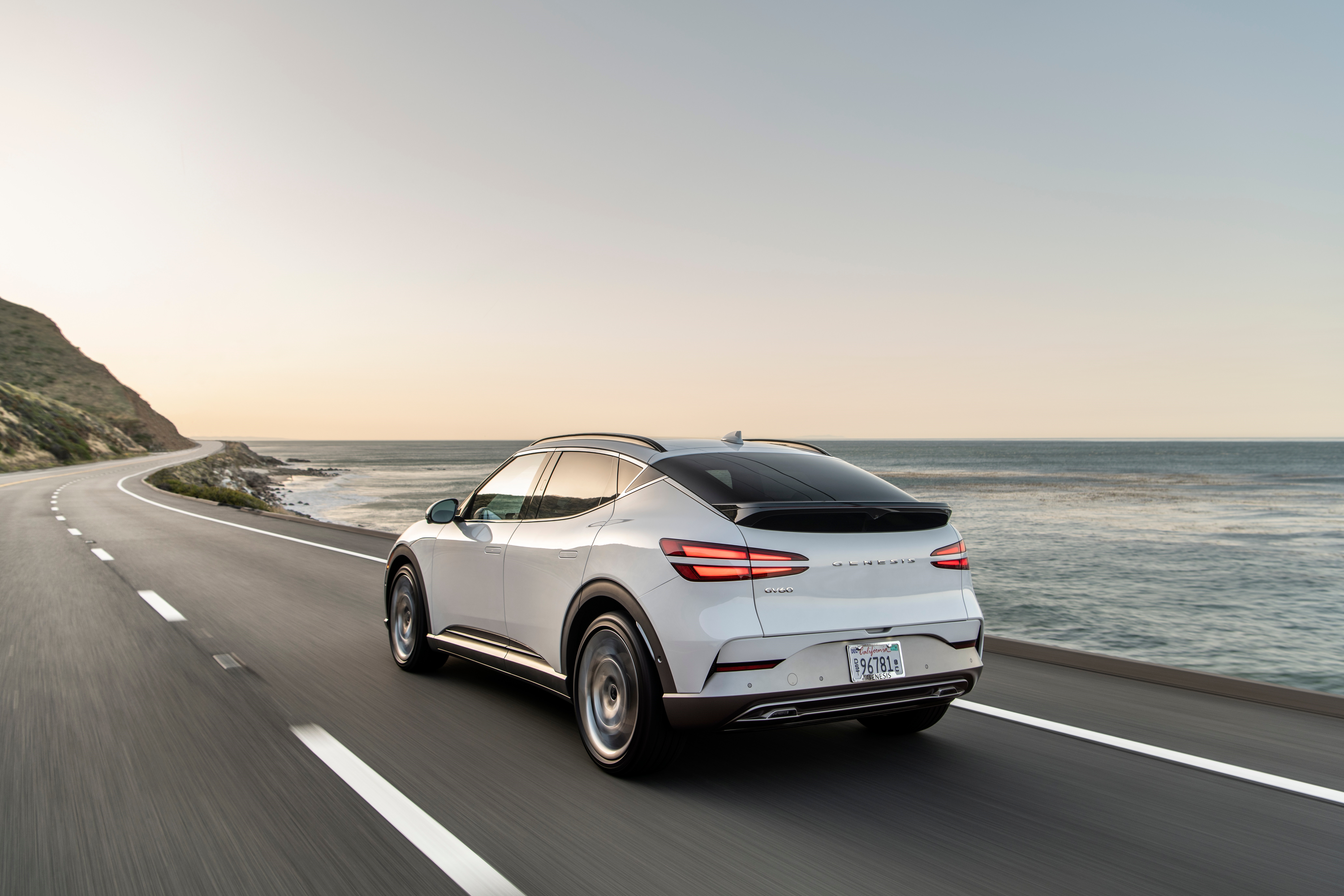 White Genesis GV60 driving on coastal highway HD Wallpaper