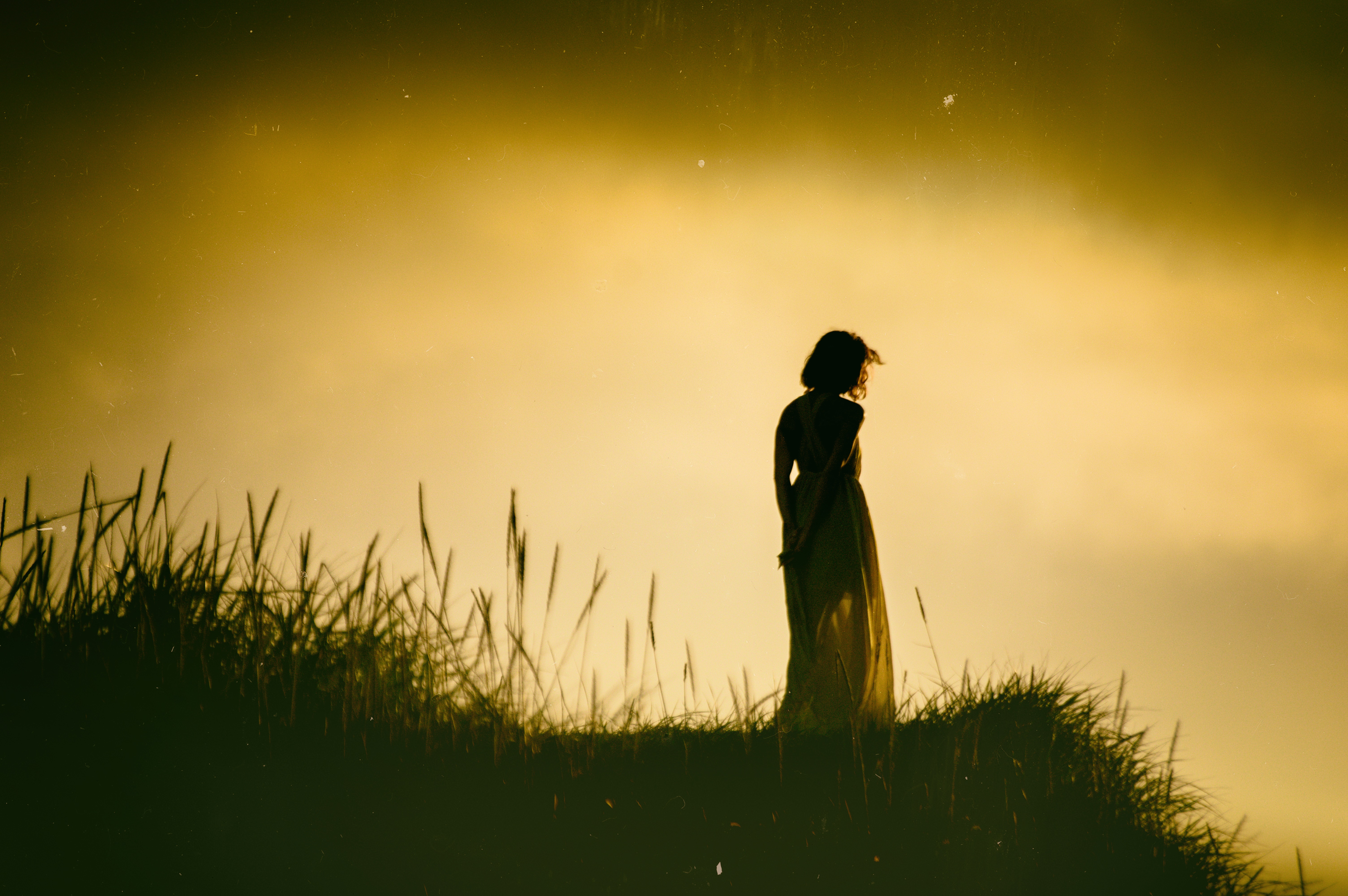 Girl in a dress standing on a grassy hill at sunset HD Wallpaper
