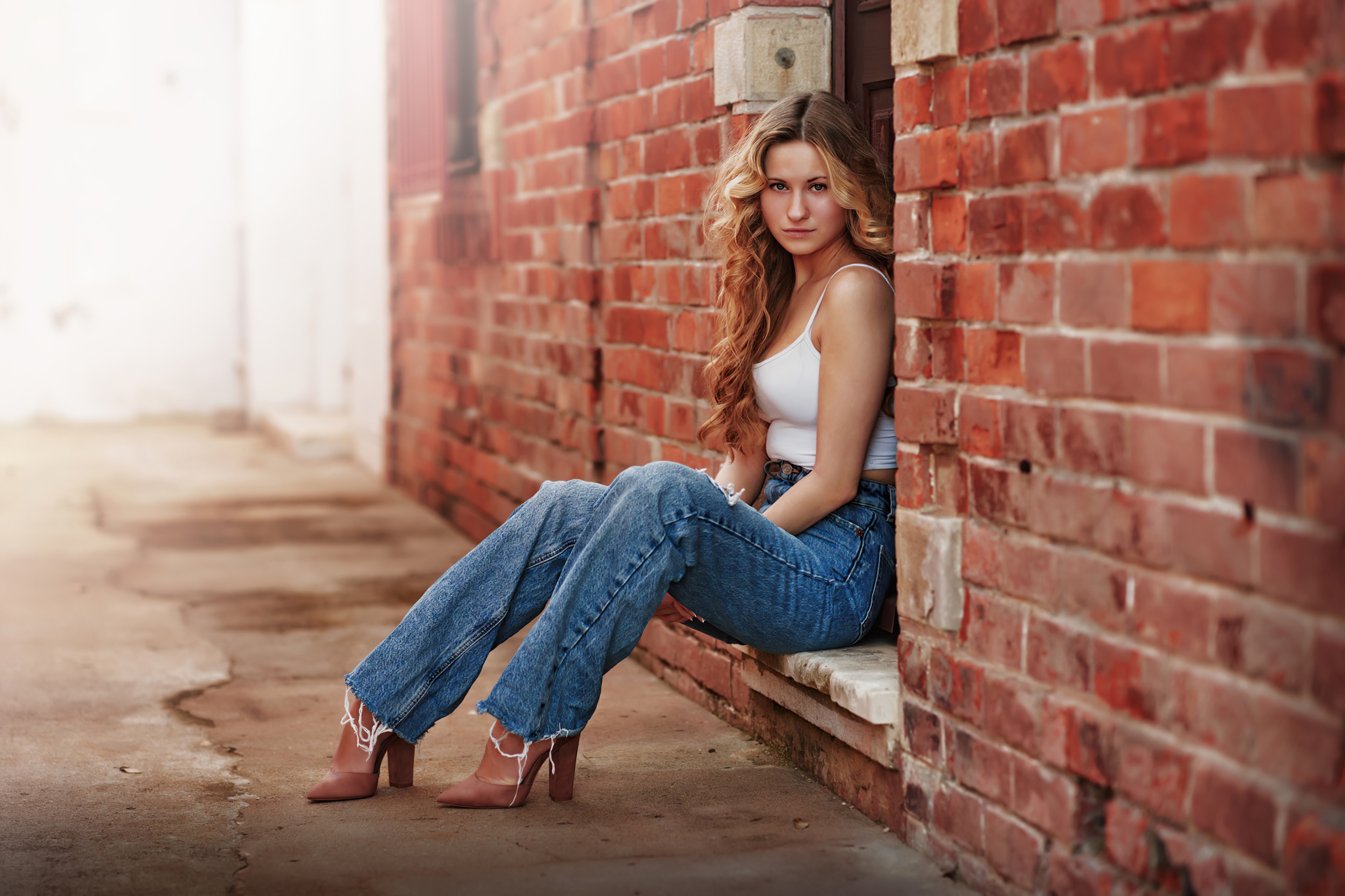 Blonde girl in jeans and white top sitting by brick wall HD Wallpaper