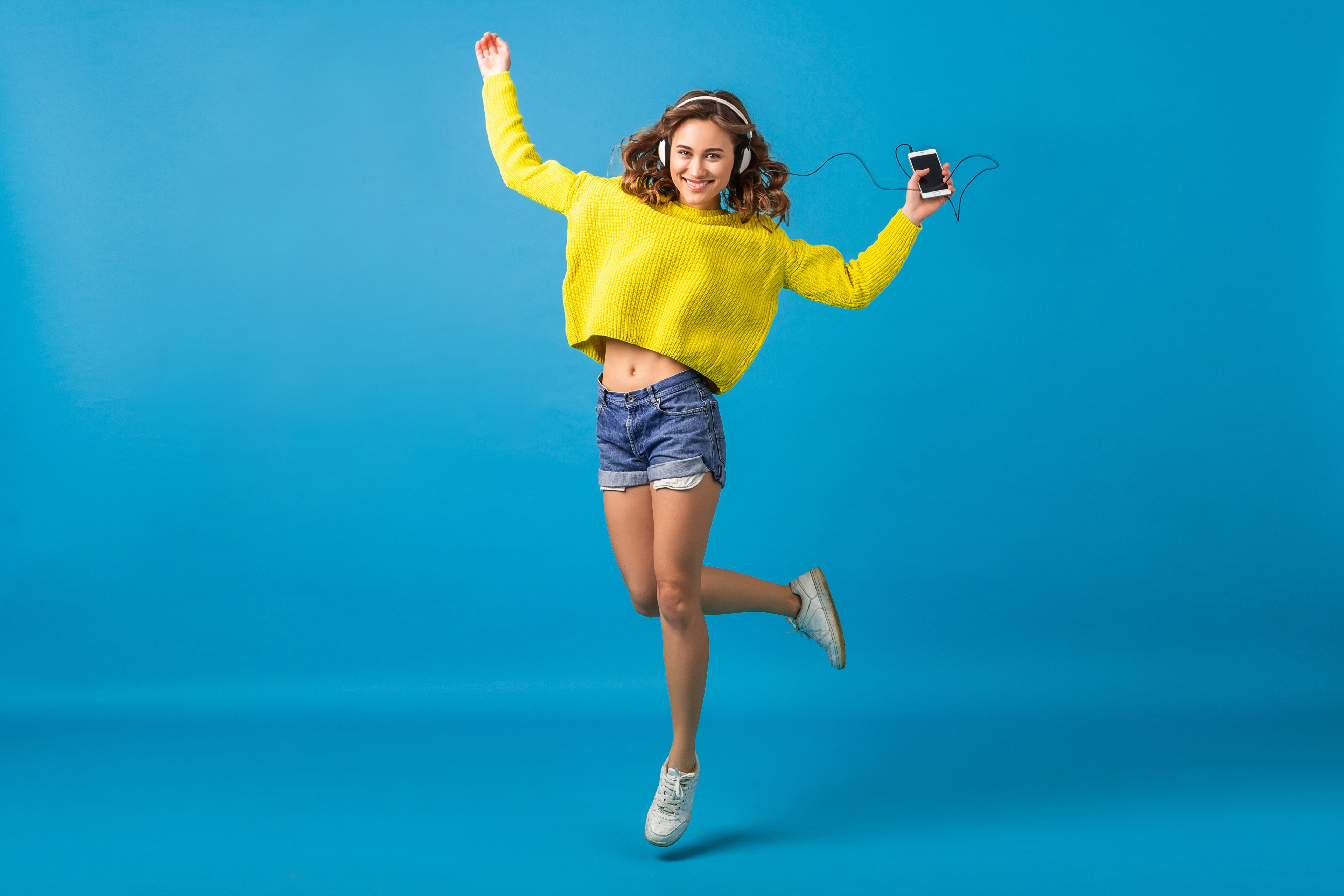 Woman in yellow sweater and denim shorts jumping with headphones and smartphone HD Wallpaper