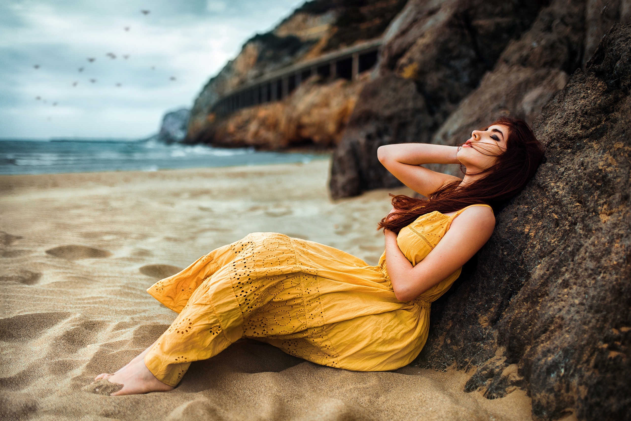Woman in yellow dress sitting on a beach by rocks HD Wallpaper