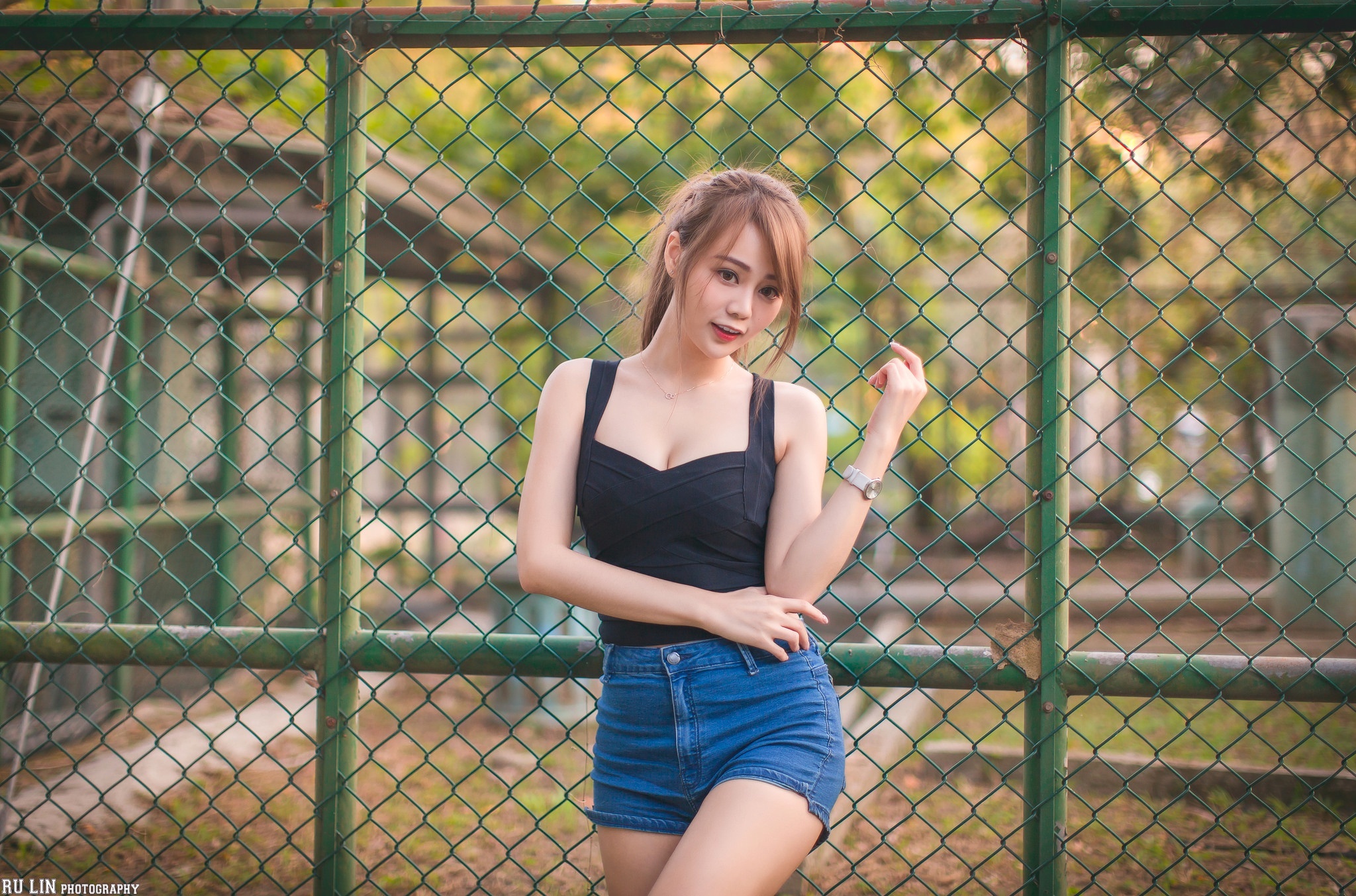Asian woman in black top and denim shorts leaning on fence HD Wallpaper