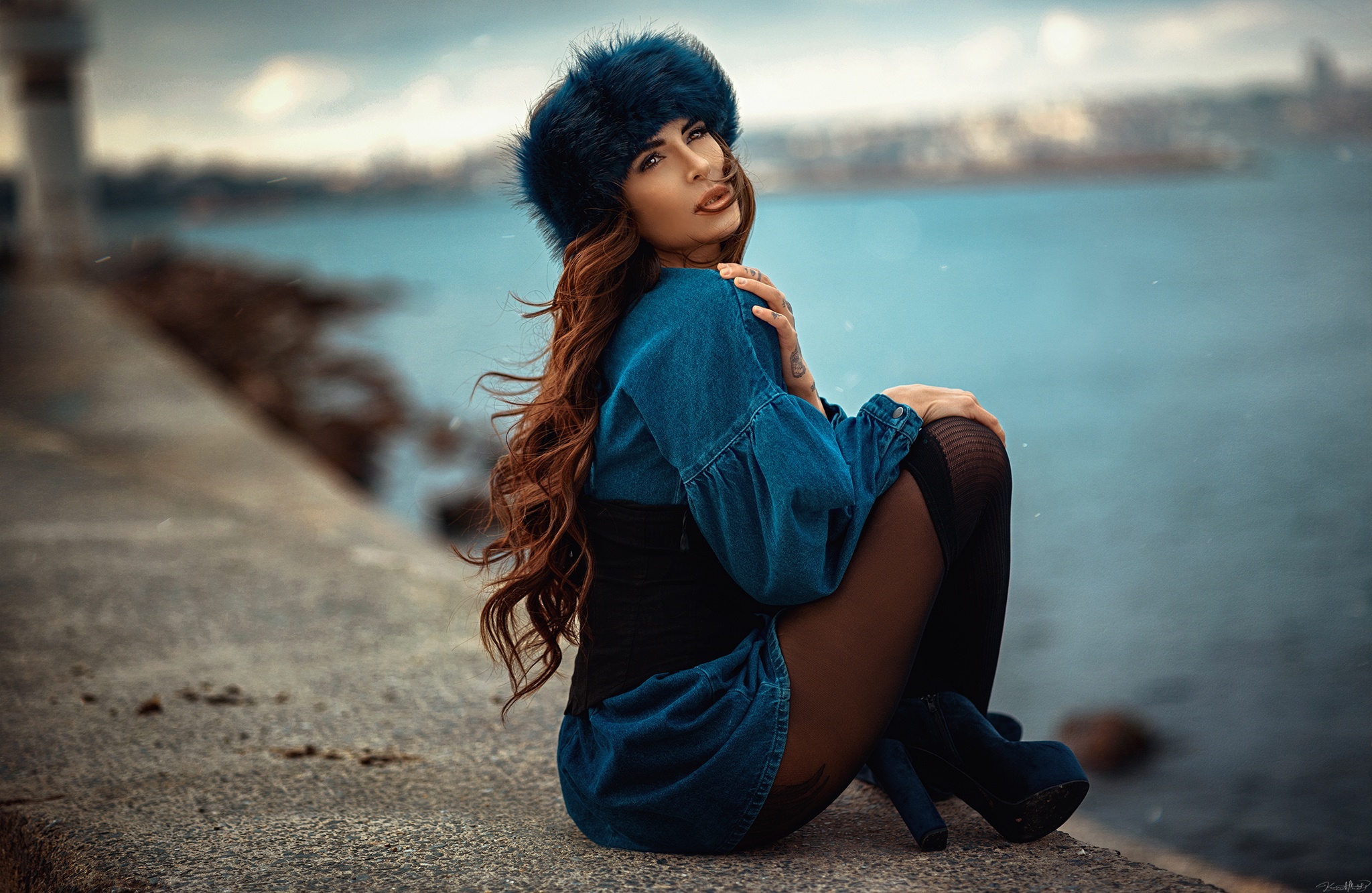 Woman in denim dress and fur hat sitting by the water HD Wallpaper