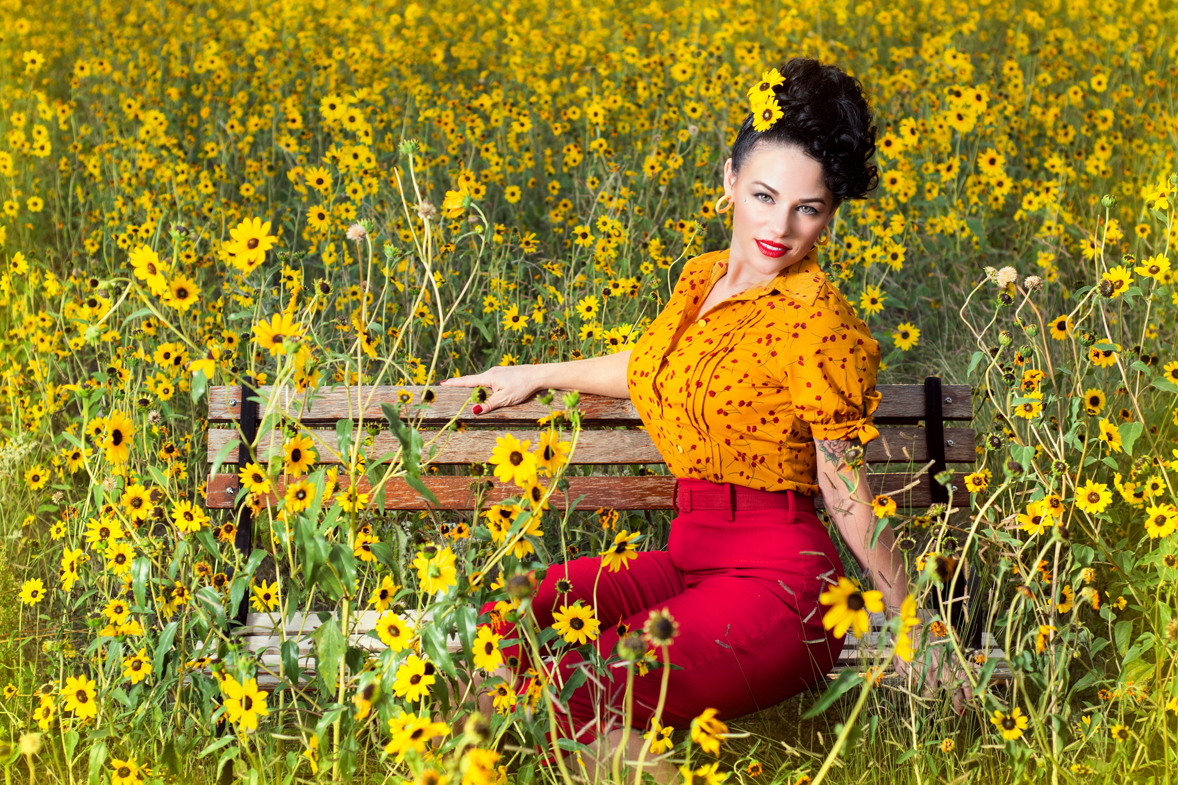 Woman in yellow blouse and red pants sitting on a bench in a field of yellow flowers HD Wallpaper