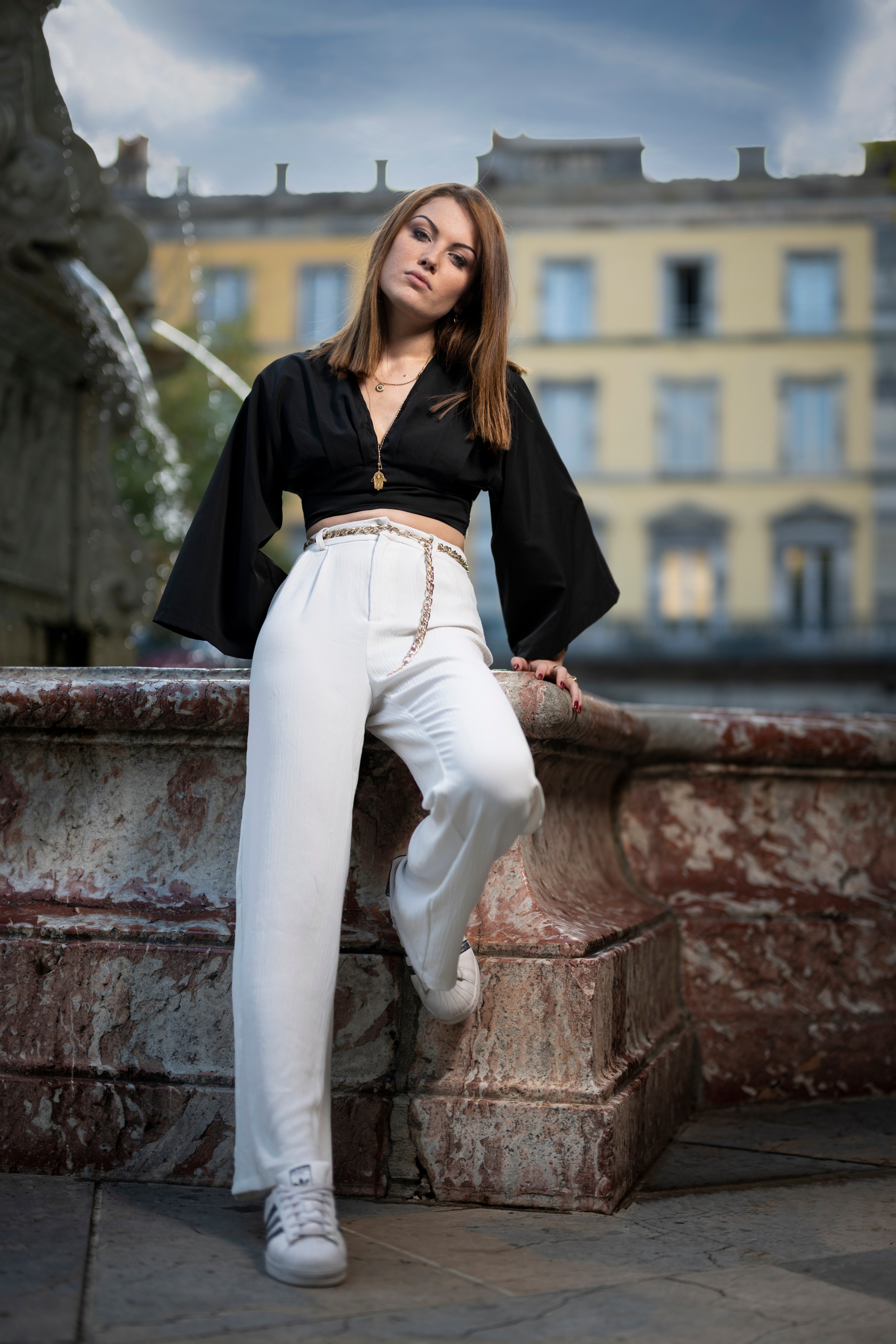 Woman in black blouse and white trousers posing by fountain HD Phone Wallpaper