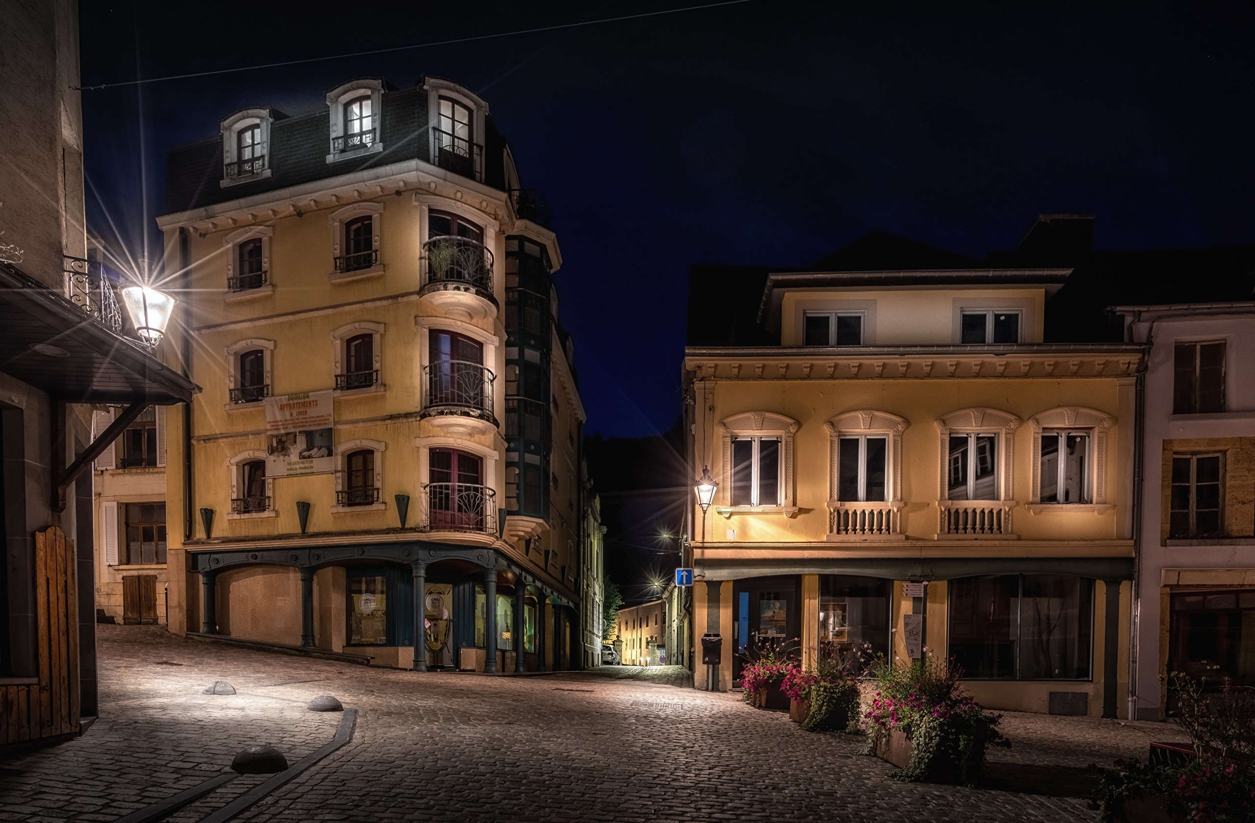 Cobblestone street at night with illuminated buildings and streetlights HD Wallpaper