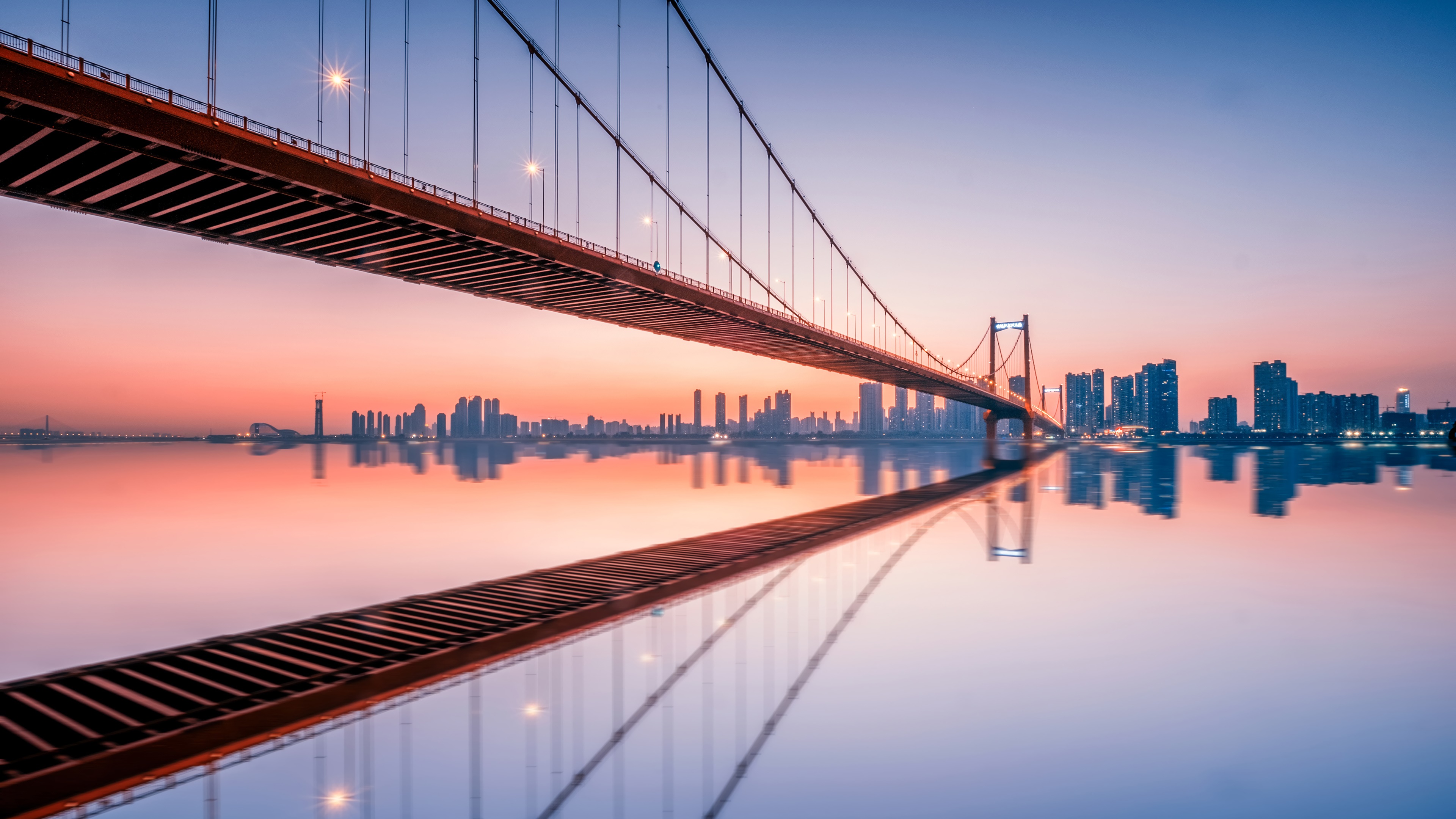 Suspension bridge over a river with city skyline at sunset 4K HD Wallpaper