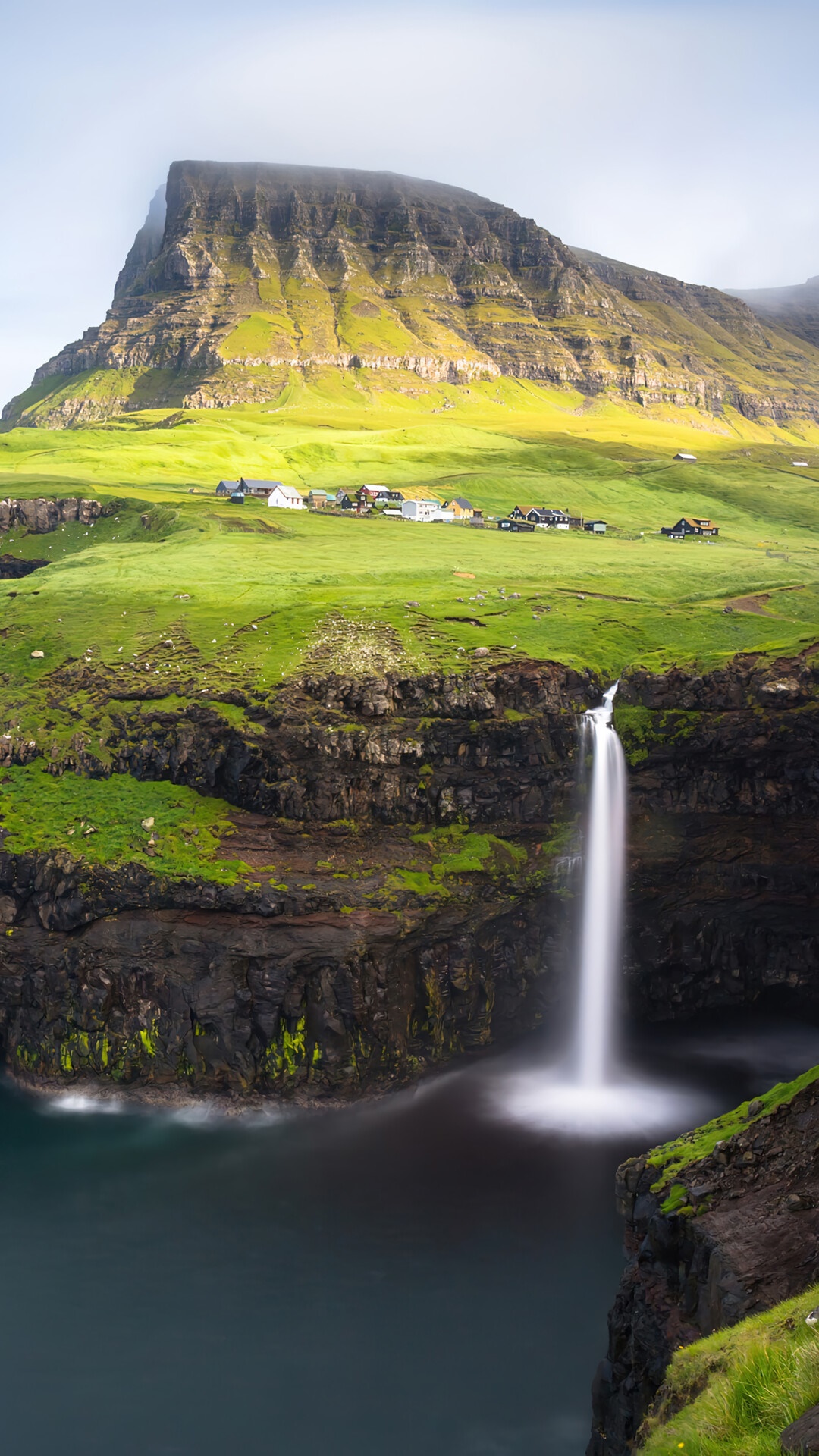 Waterfall cascading into the sea near a village on a green island HD Phone Wallpaper