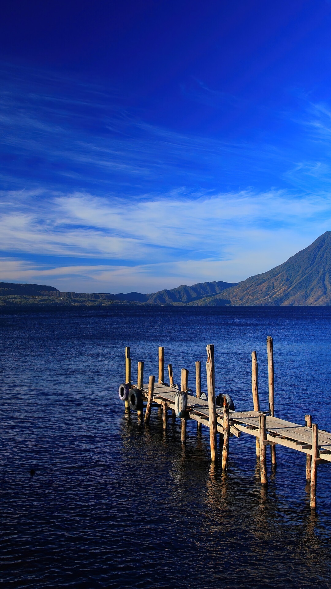 Wooden pier on a deep blue lake with mountains under a bright blue sky HD Phone Wallpaper