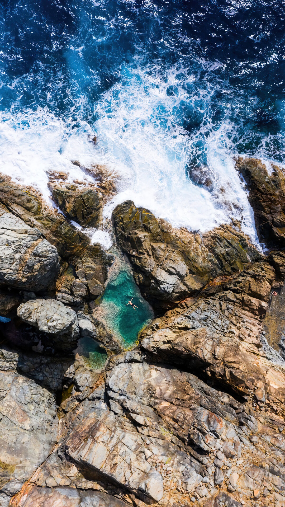 Aerial view of a person swimming in a natural ocean pool surrounded by rocks HD Phone Wallpaper