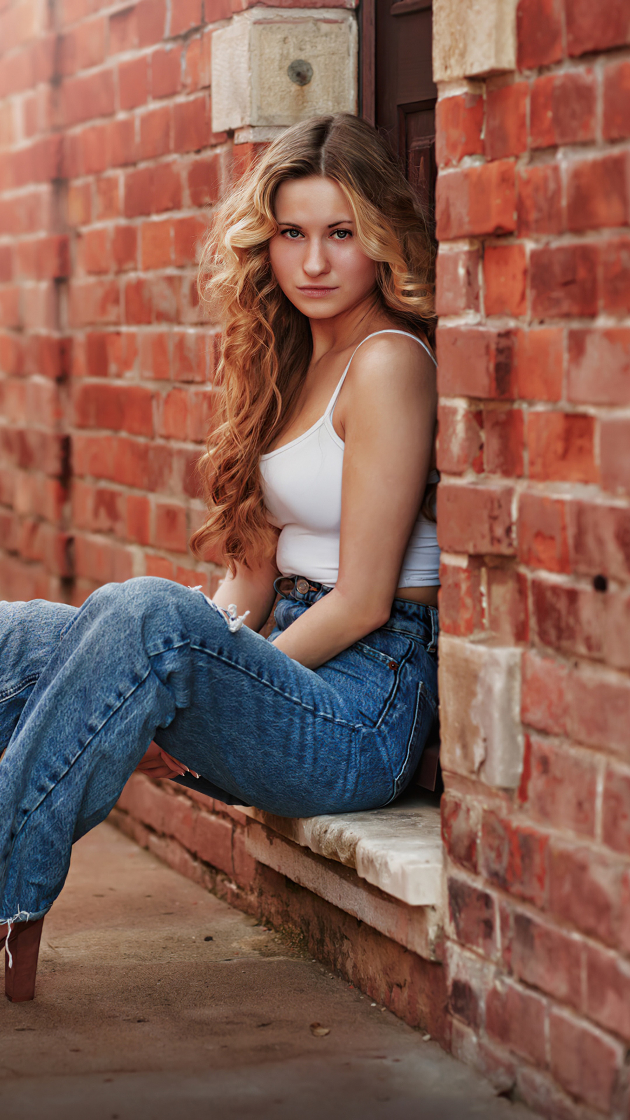 Blonde girl in white top and jeans sitting by brick wall HD Phone Wallpaper