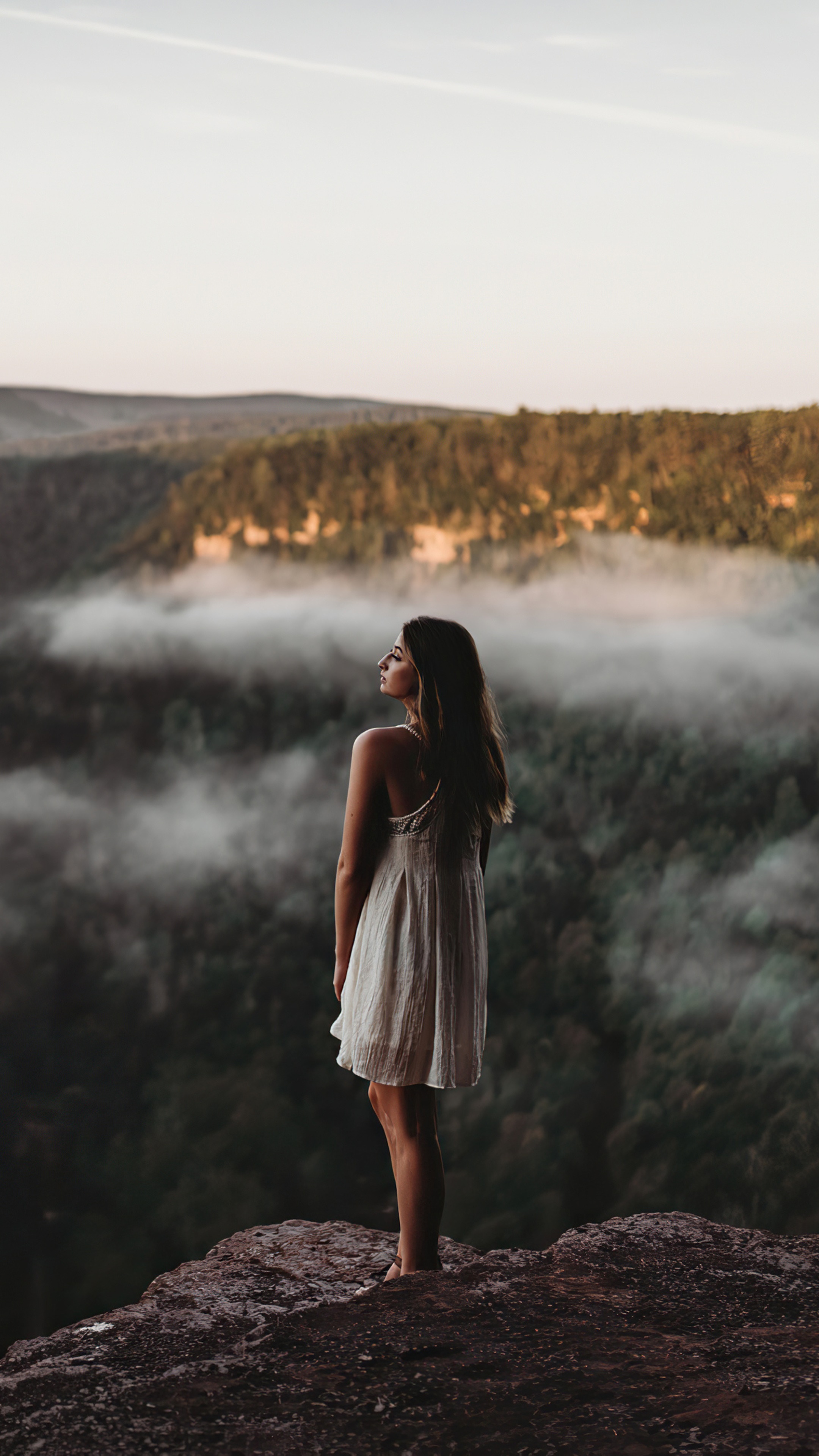 Girl in white dress on cliff edge overlooking foggy forest HD Phone Wallpaper
