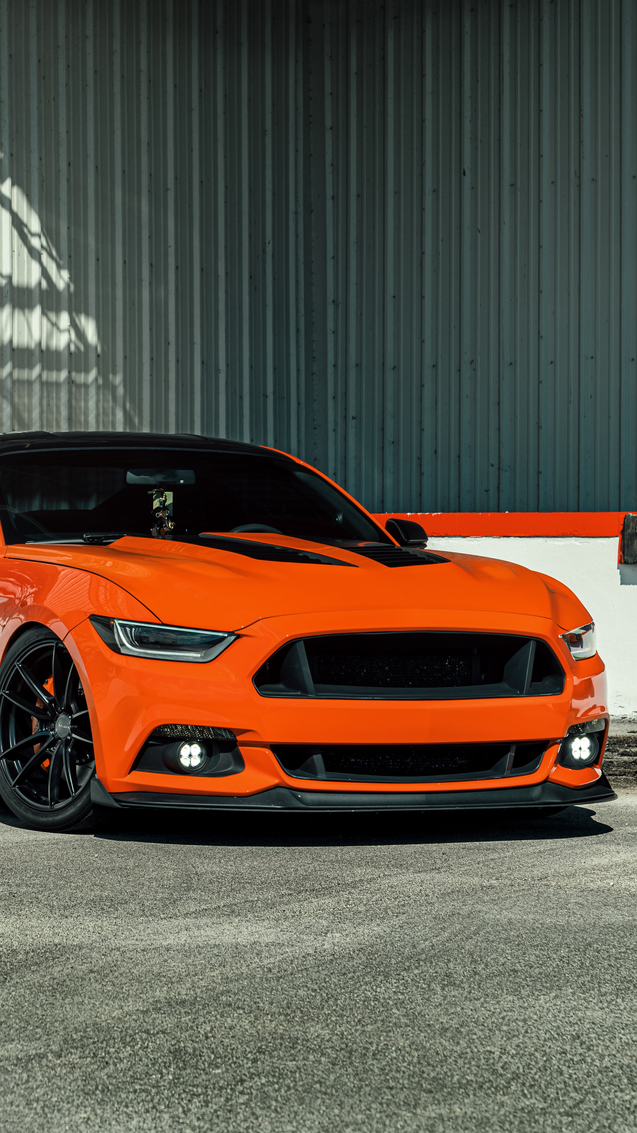 Orange Ford Mustang parked in front of a corrugated metal wall HD Phone Wallpaper