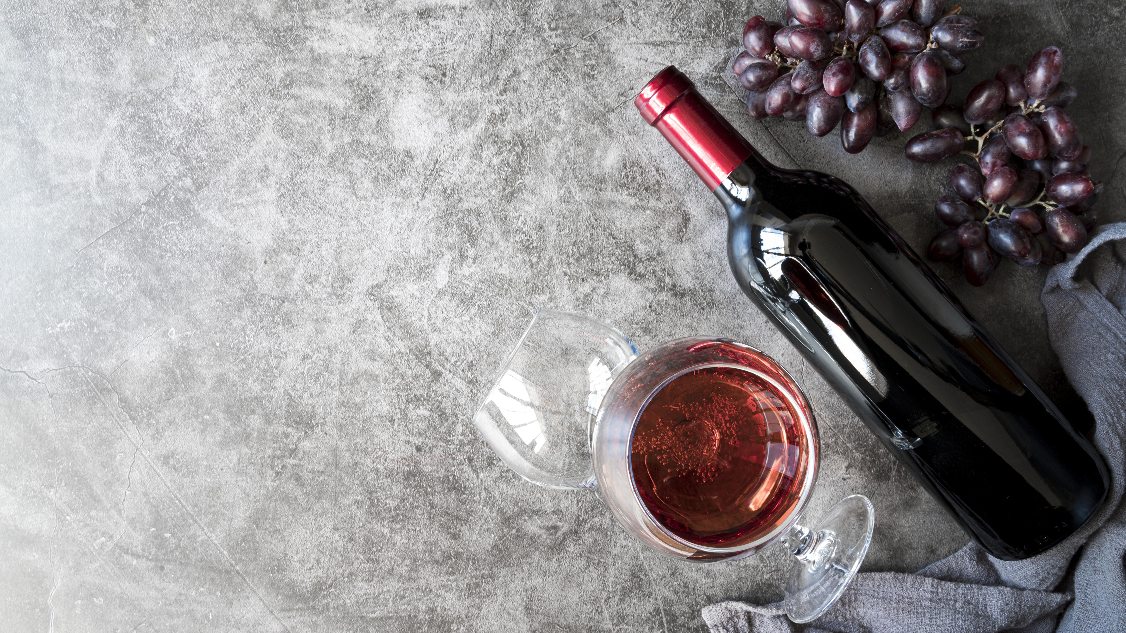 Red wine bottle, glass, and grapes on a textured surface HD Wallpaper