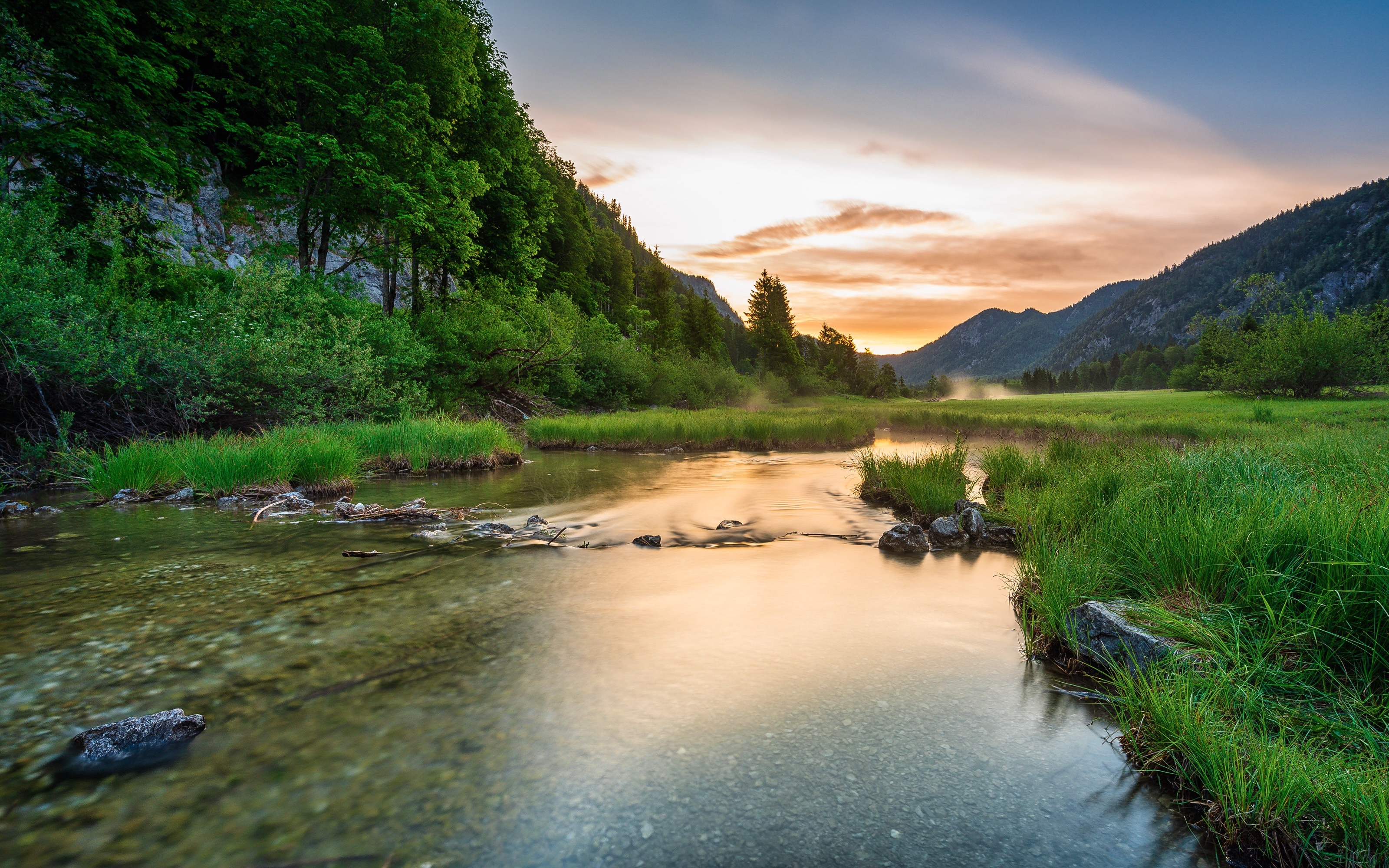 River flowing through a grassy meadow at sunset with mountains HD Wallpaper