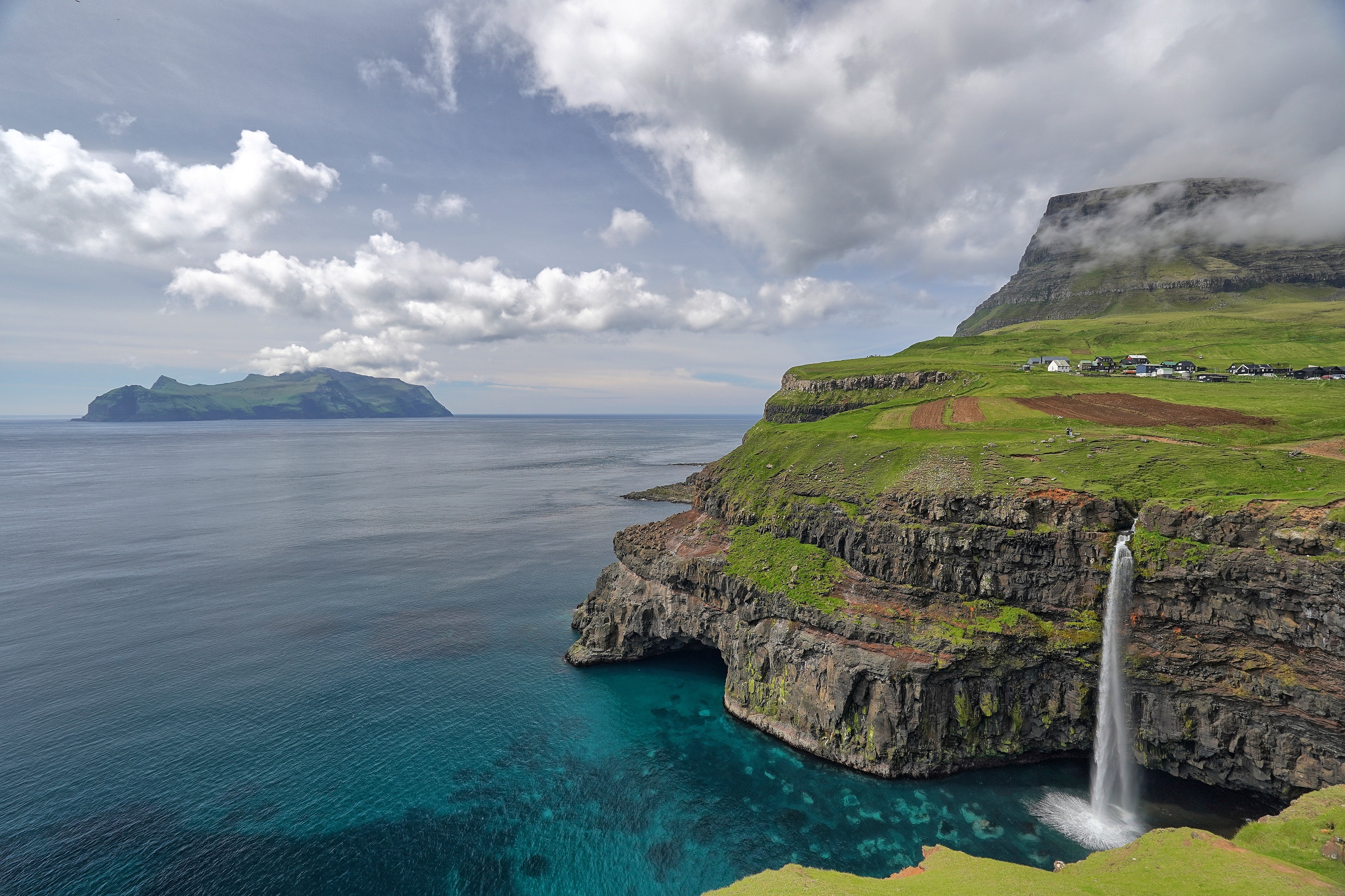 Waterfall cascading into the ocean near a village on a cliffside HD Wallpaper