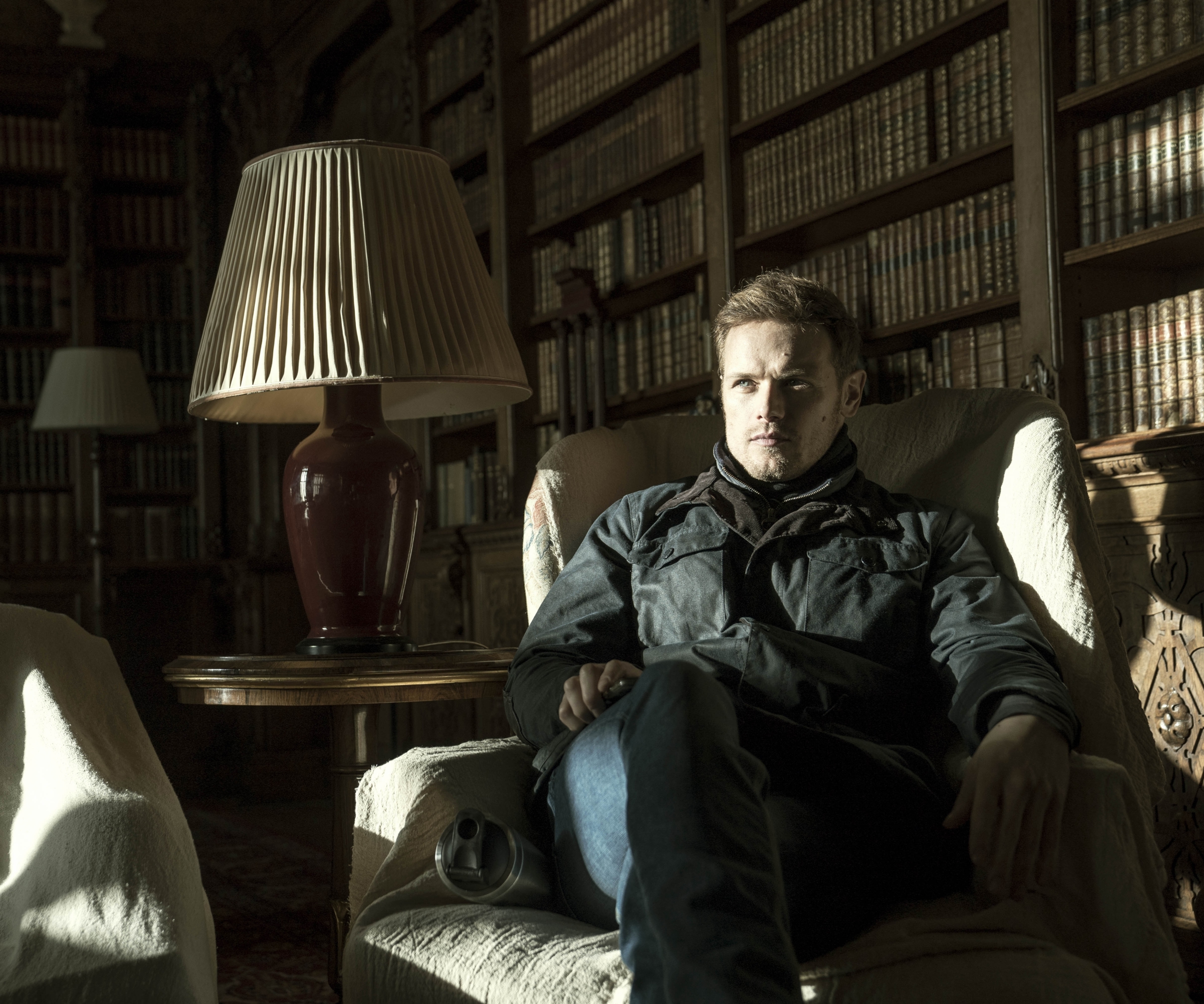 Sam Heughan sitting in a sunlit library HD Wallpaper