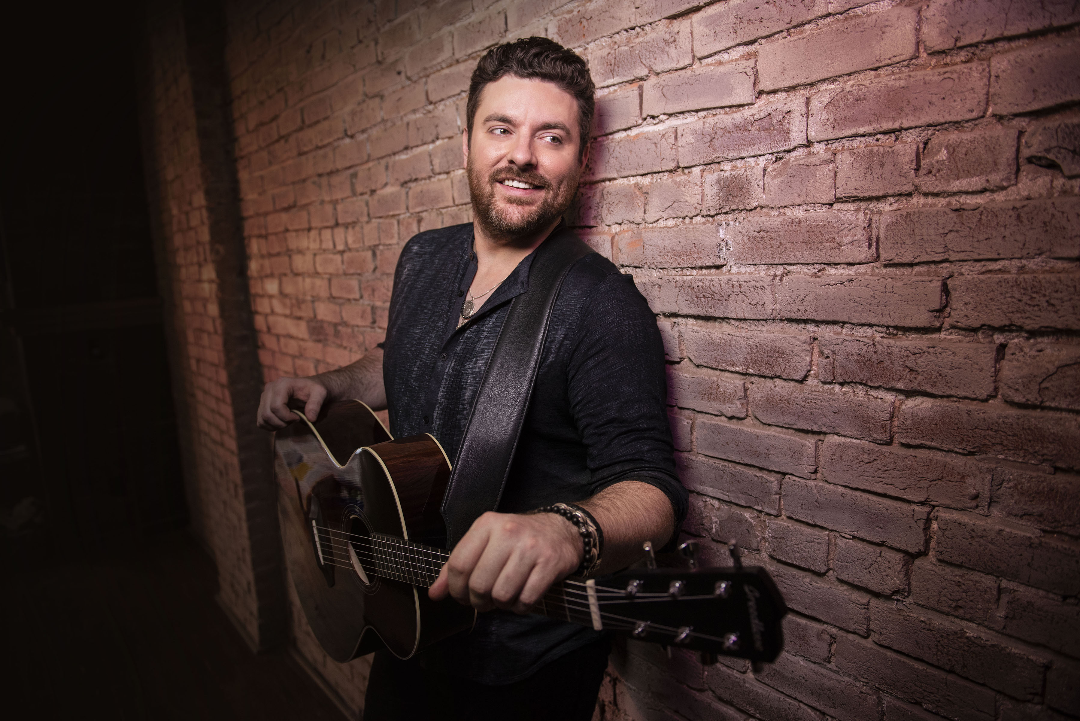 Chris Young smiling with acoustic guitar against brick wall HD Wallpaper