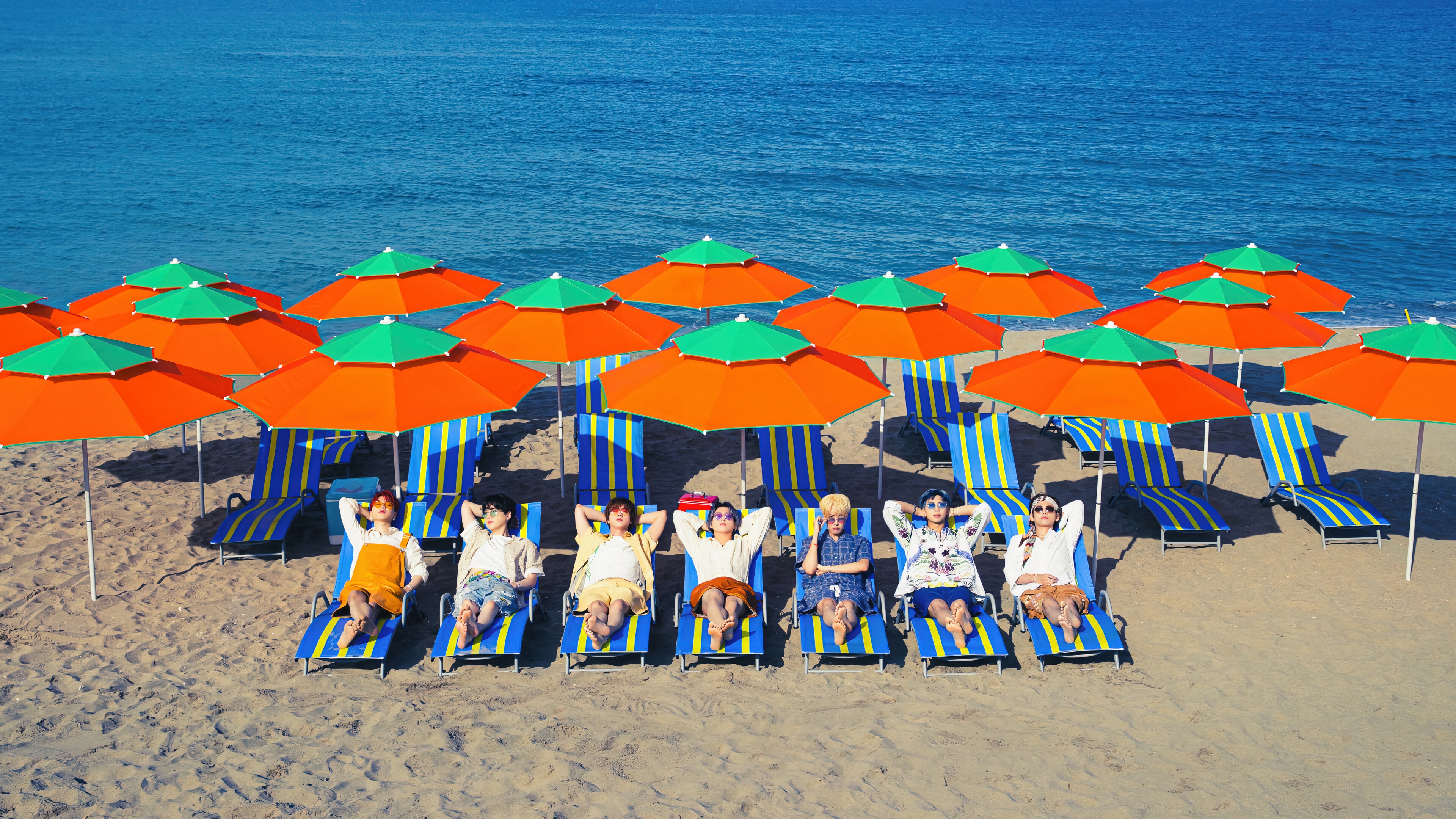 BTS members relaxing on a beach with umbrellas HD Wallpaper