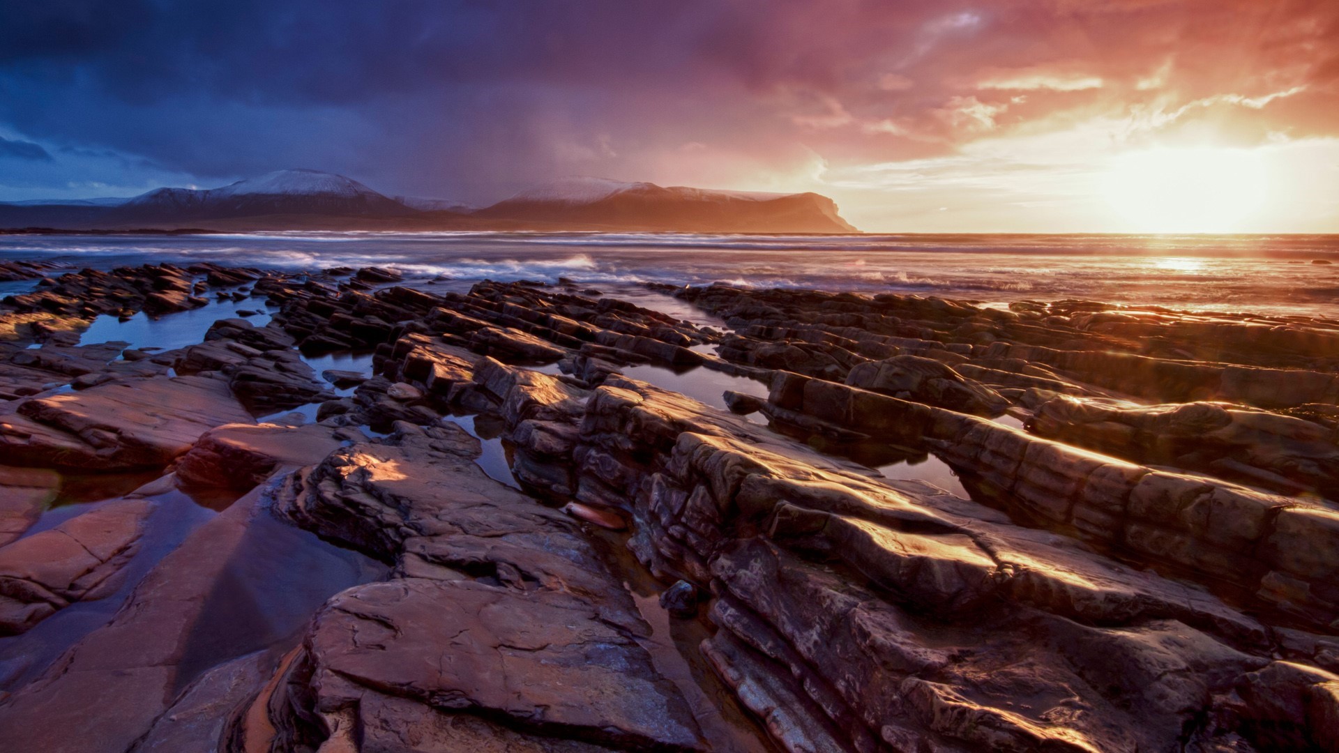 Rocky coastline at sunset with mountains in the distance HD Wallpaper