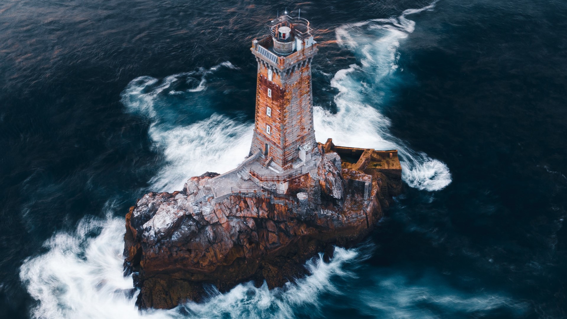 Lighthouse on rocky island surrounded by turbulent water HD Wallpaper