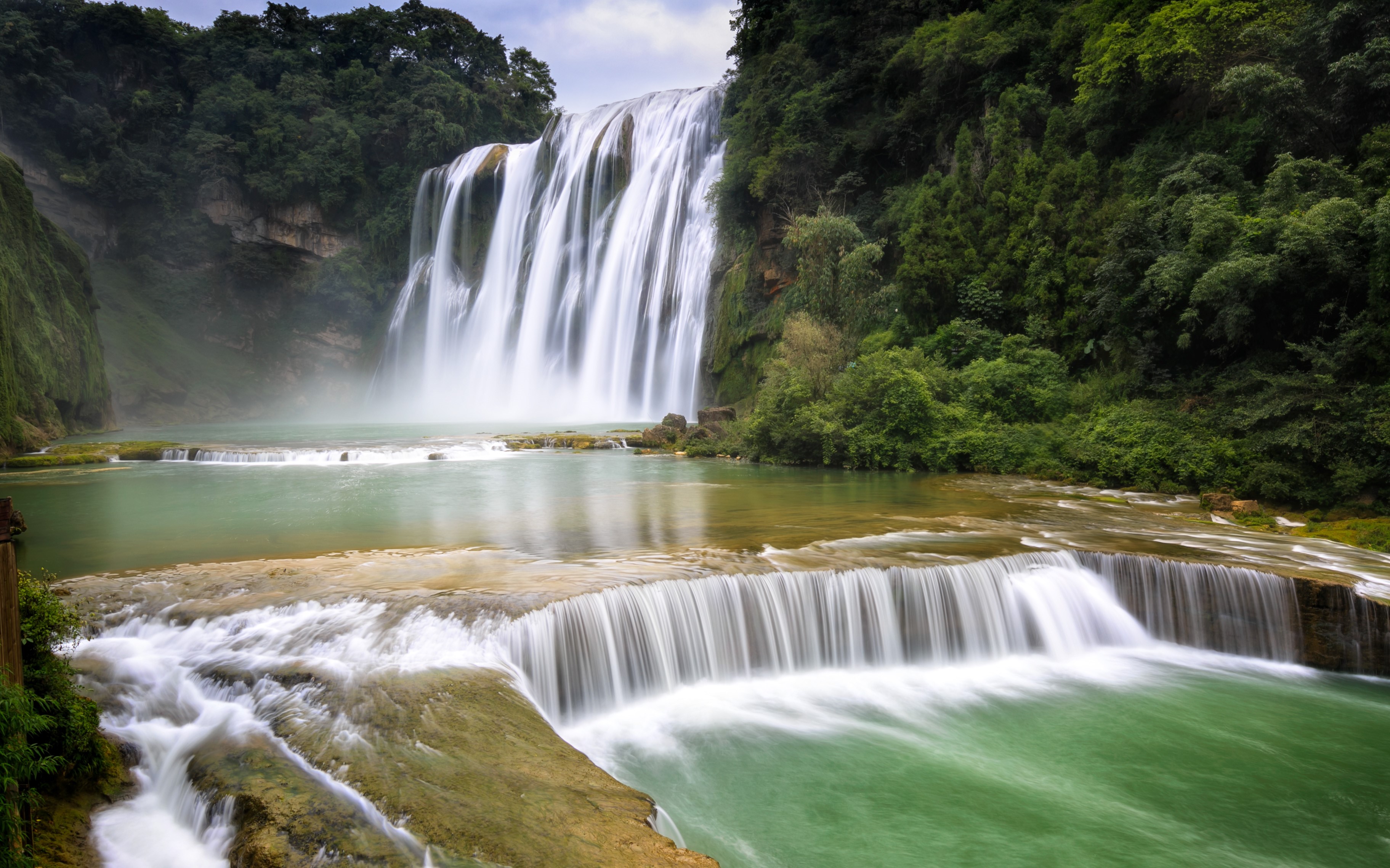 A wide waterfall cascades into a turquoise pool surrounded by lush green trees. HD Wallpaper