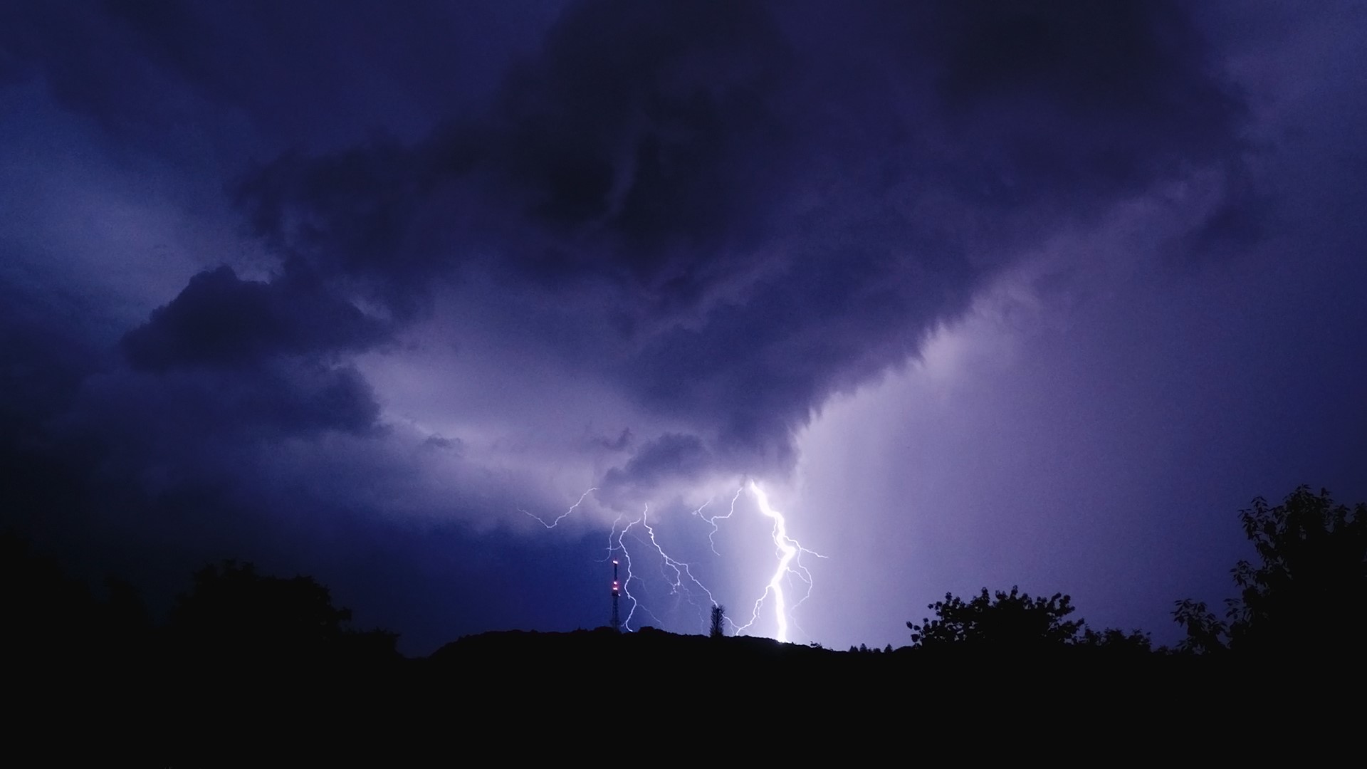 Lightning strikes a radio tower during a nighttime storm over hills HD Wallpaper