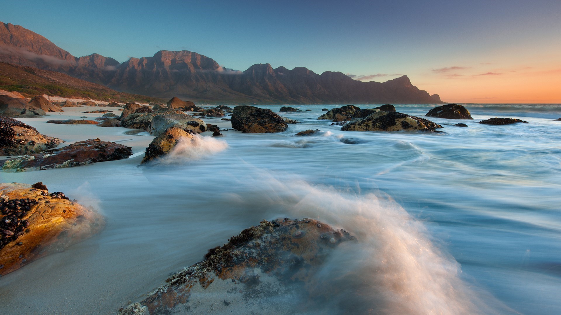 Long exposure of waves crashing on a rocky beach at sunset with mountains in the background HD Wallpaper