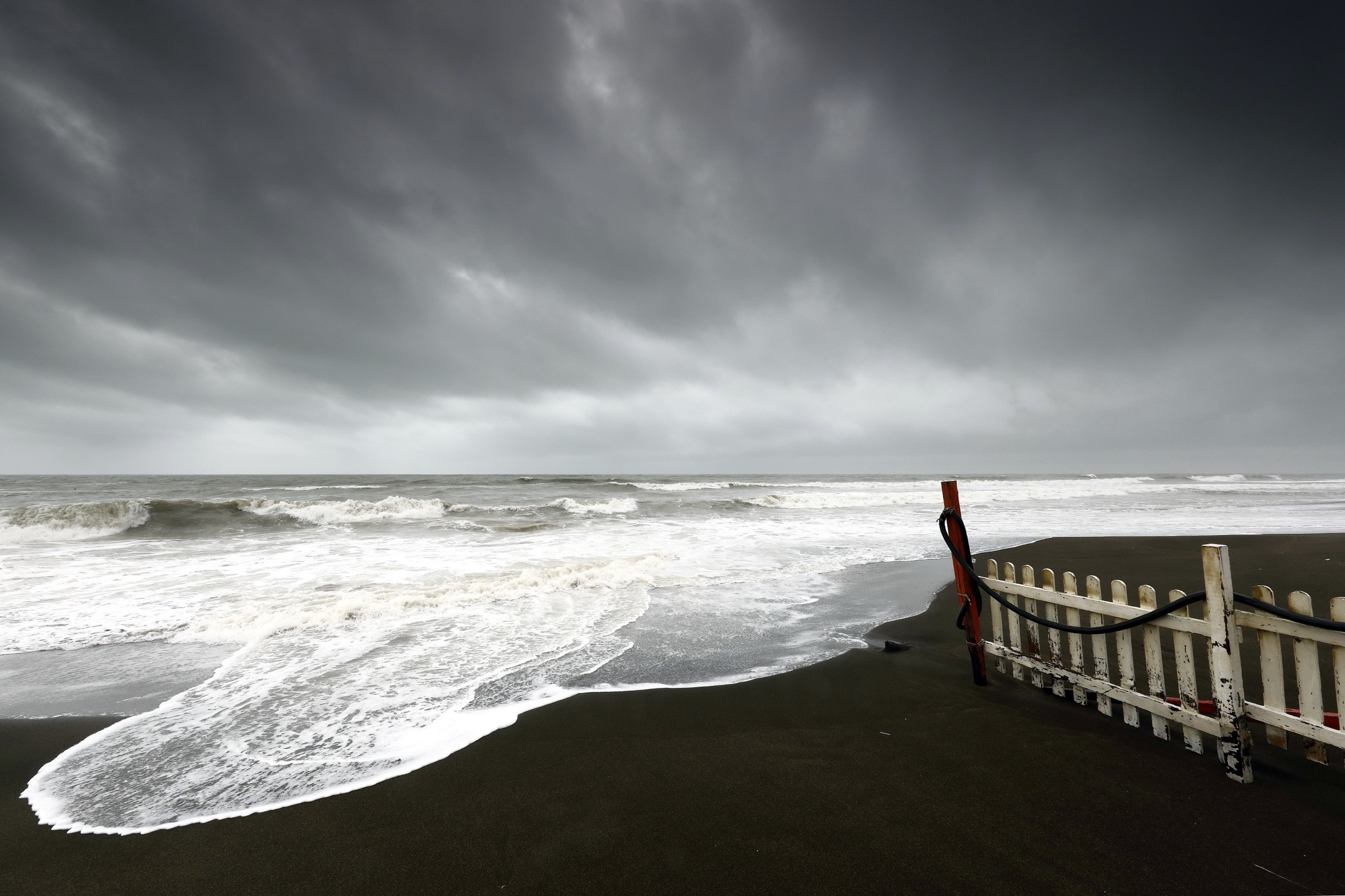 Overcast sky over a dark sand beach with waves and a white fence HD Wallpaper