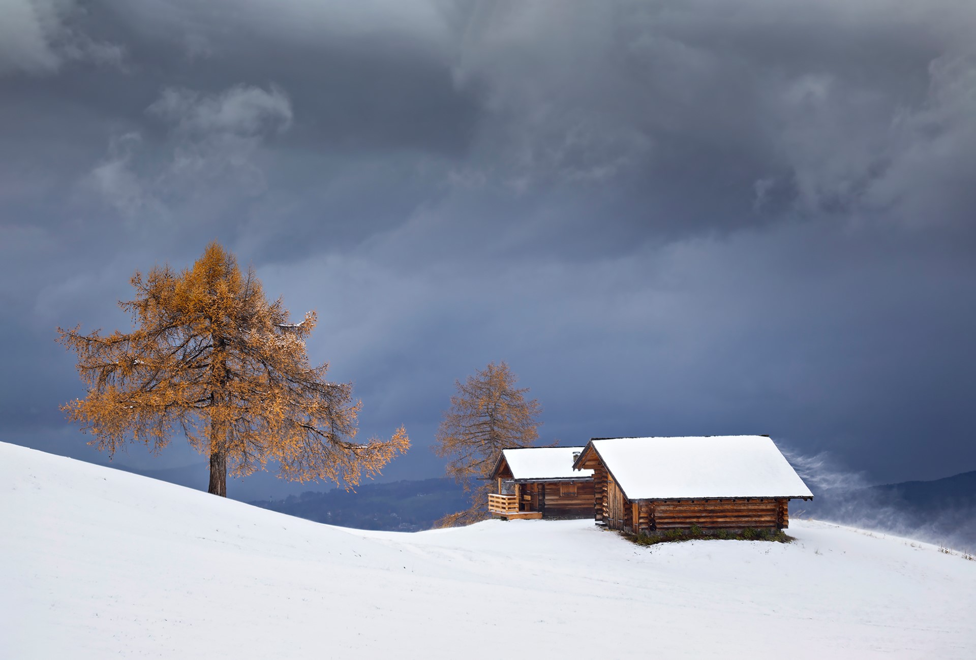 Wooden huts and a golden tree in a snowy landscape under a stormy sky HD Wallpaper