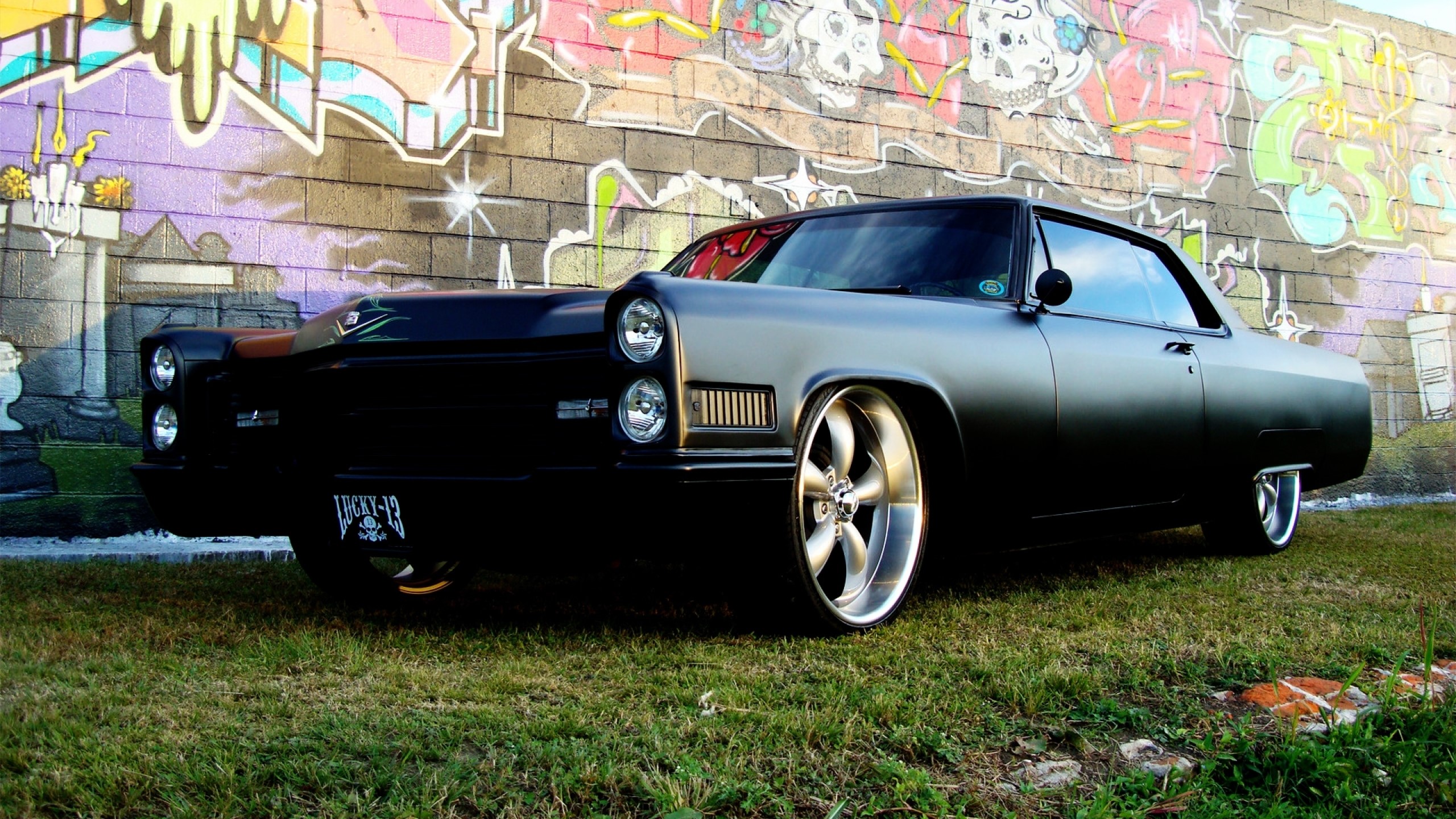 Matte black custom Cadillac parked in front of graffiti wall HD Wallpaper