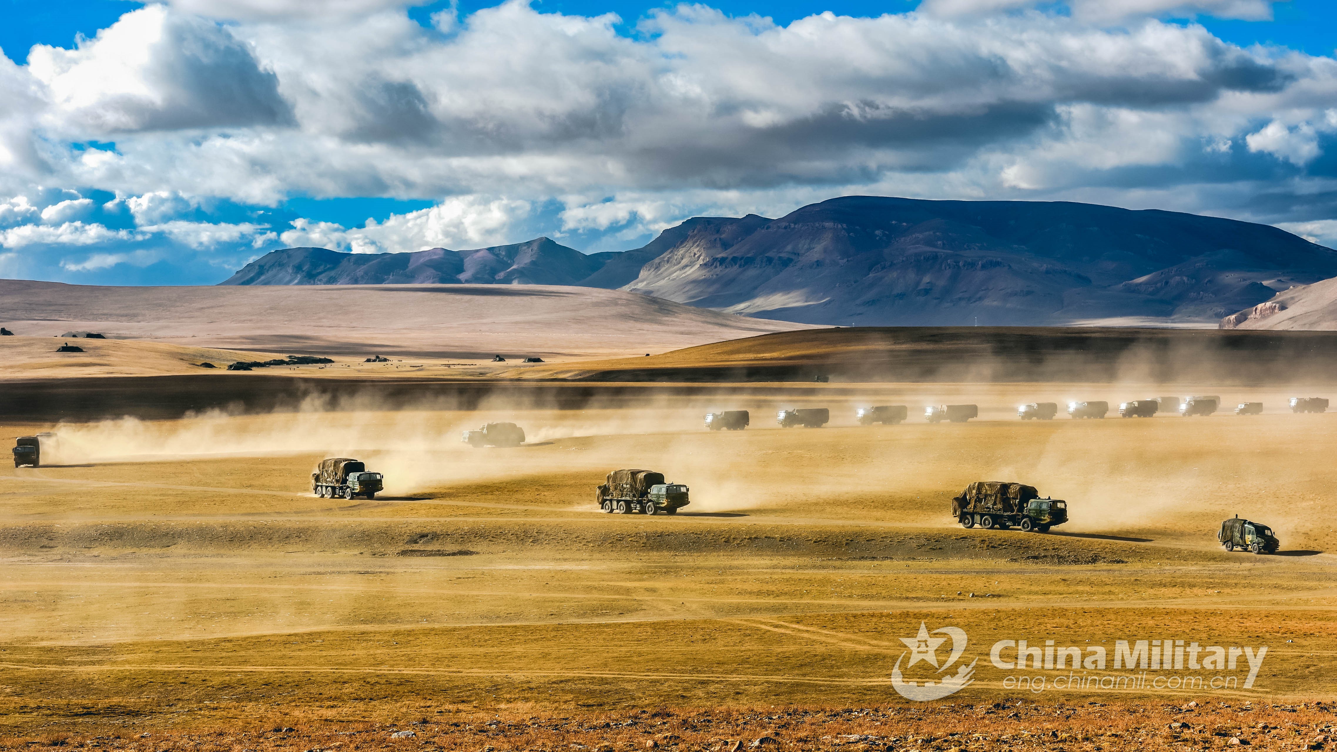 Military vehicles driving through a dusty, mountainous landscape HD Wallpaper