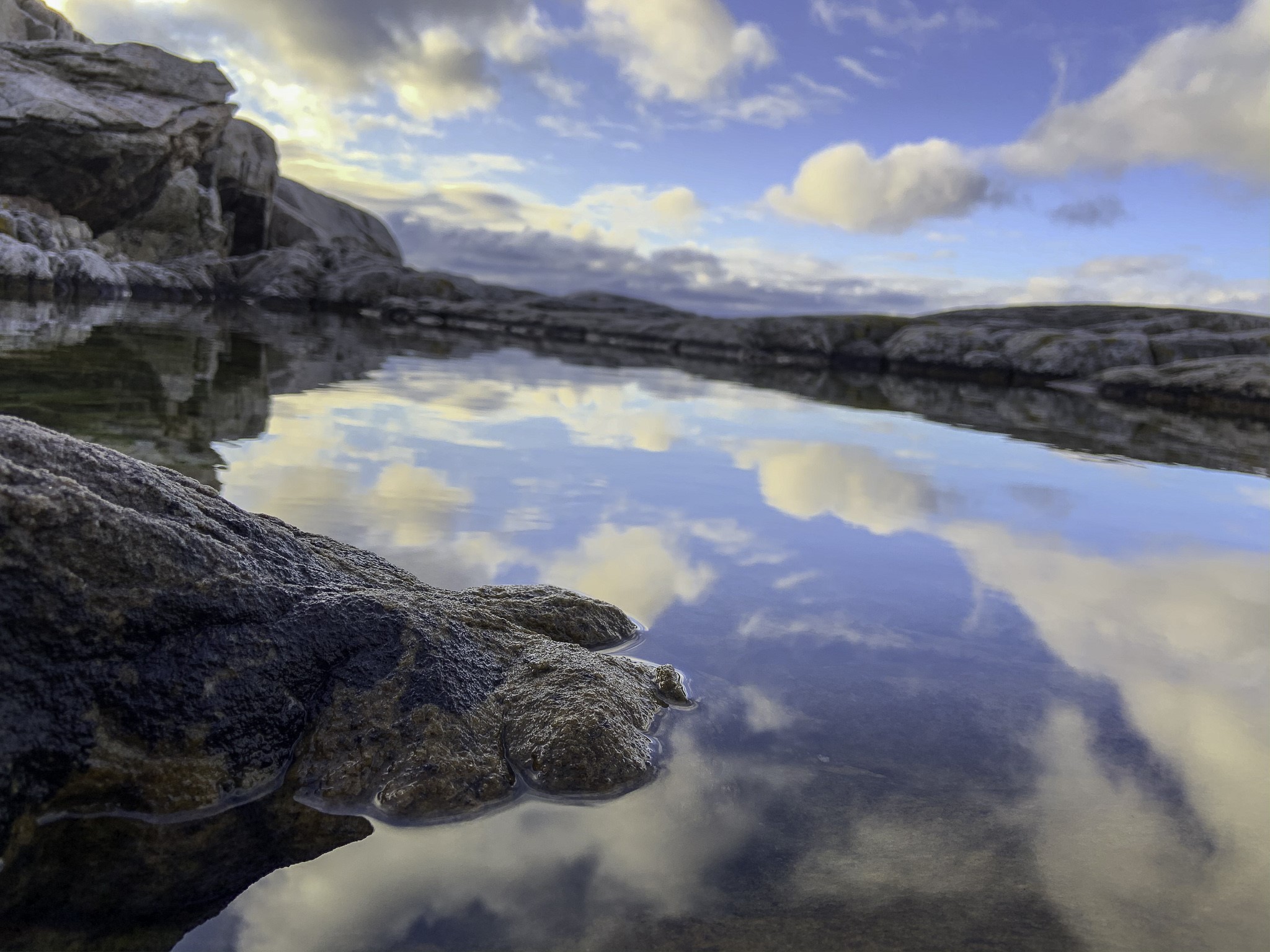 Rocky shoreline with calm water reflecting clouds HD Wallpaper