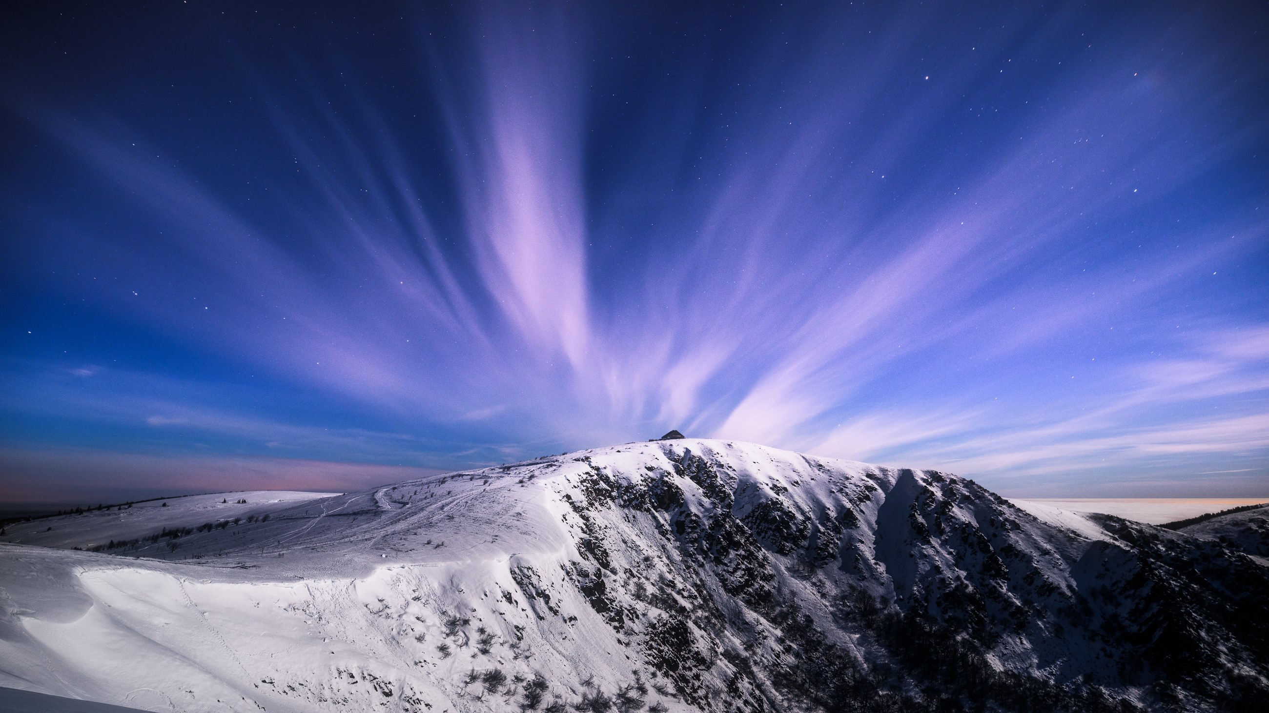 Snowy mountain peak under a starry sky with streaking clouds HD Wallpaper