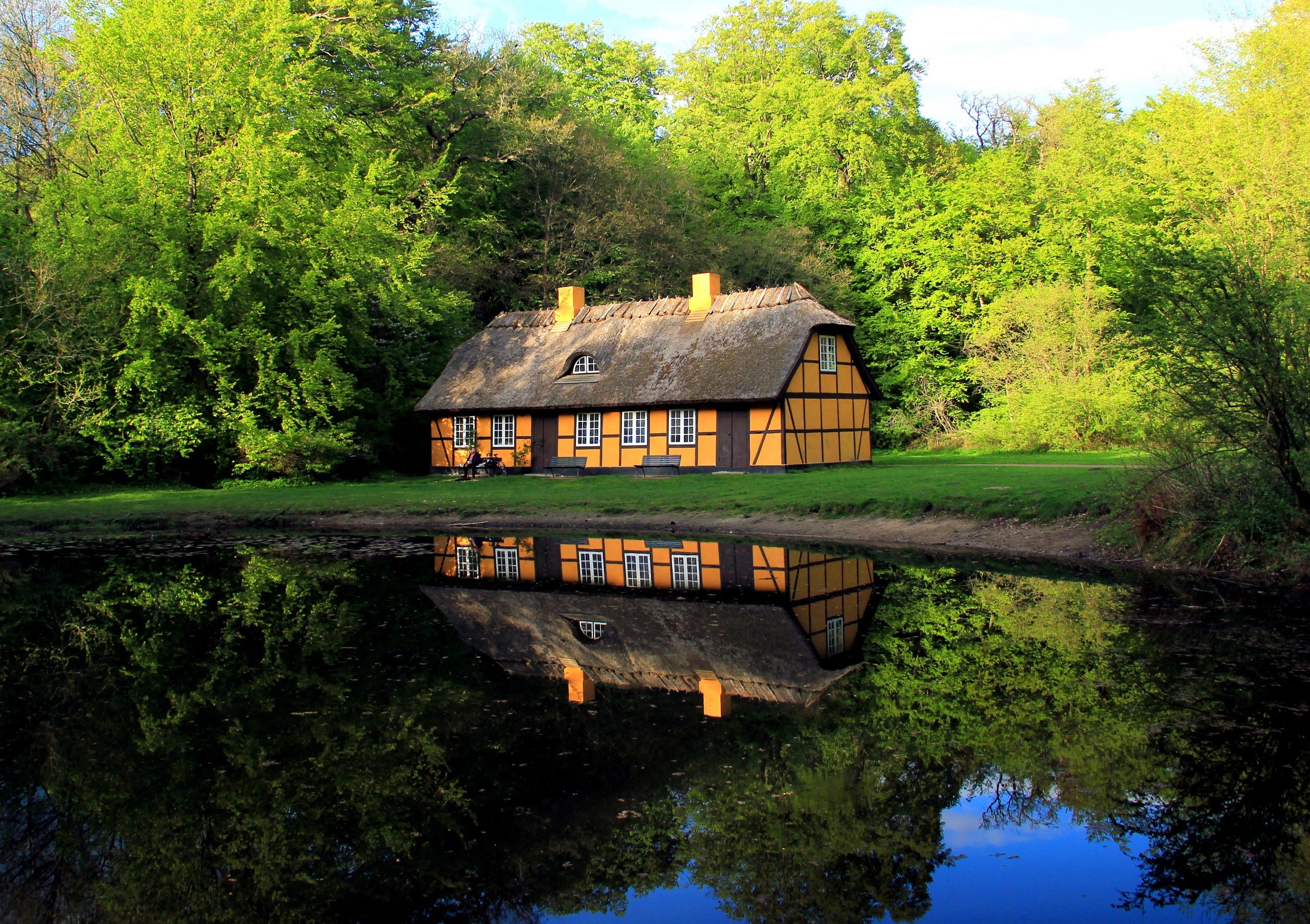 Thatched roof house reflected in a calm lake surrounded by trees HD Wallpaper