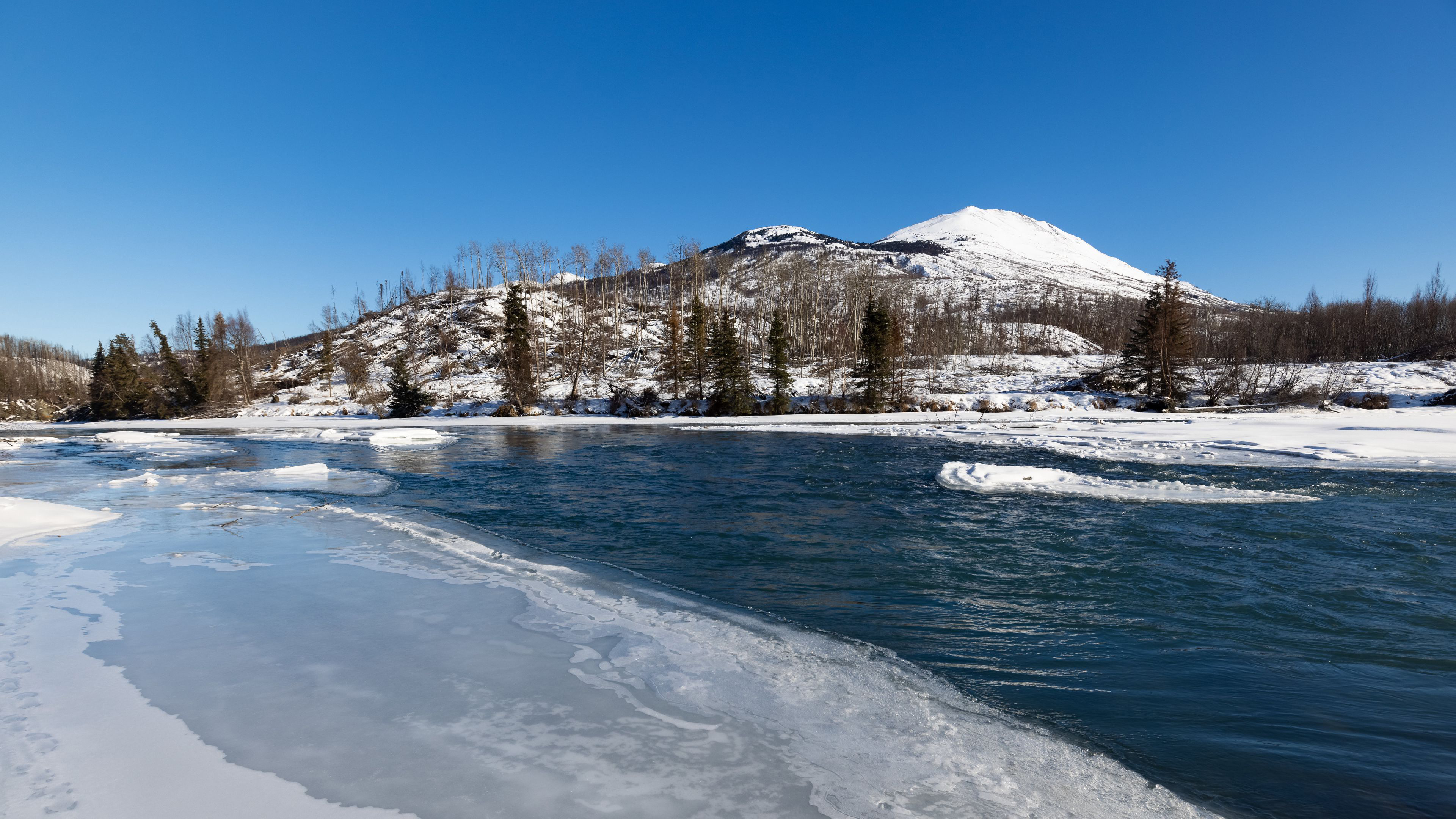 Snowy mountains and trees by a partially frozen river HD Wallpaper