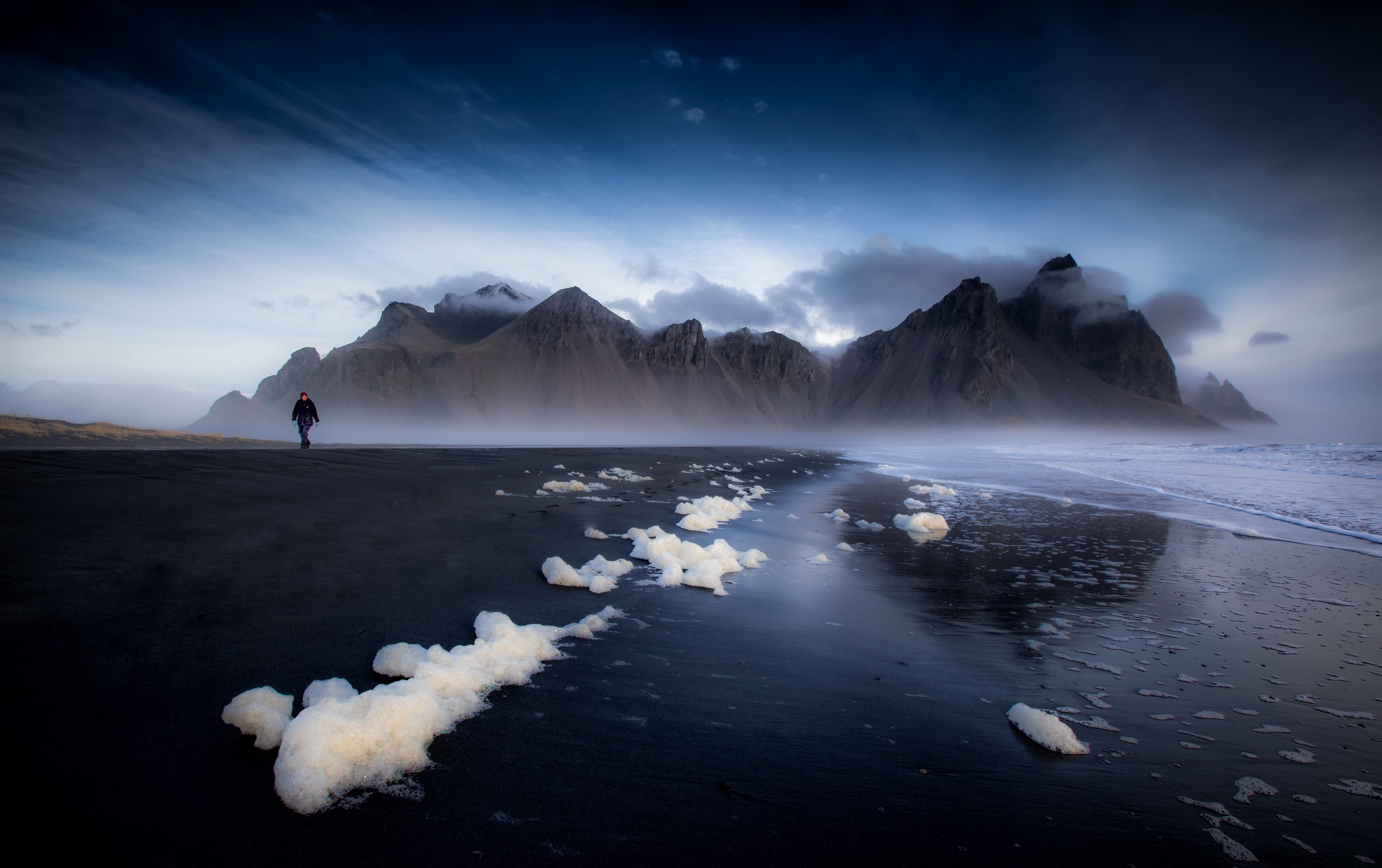 Person walking on black sand beach with mountains in Iceland HD Wallpaper