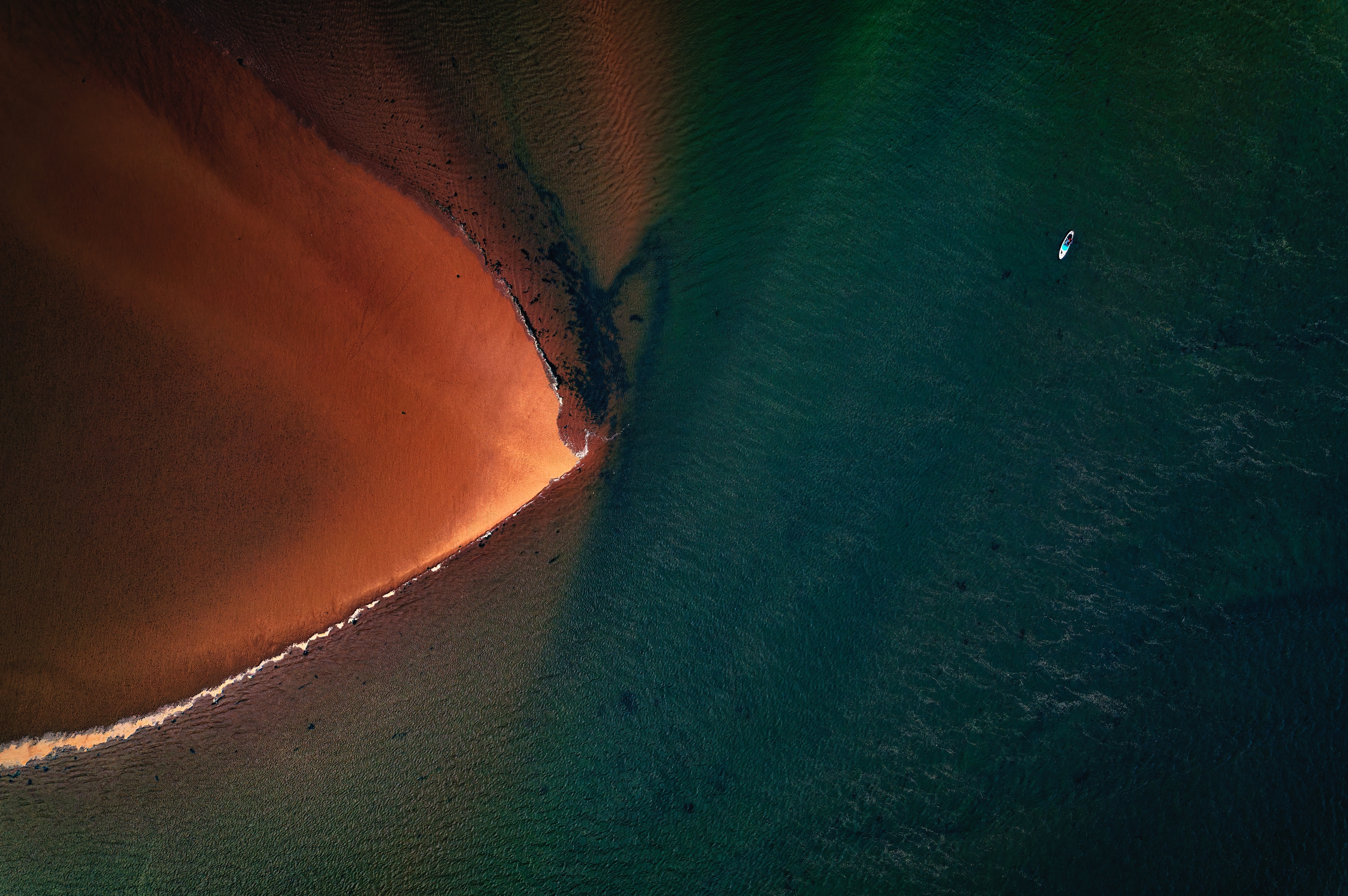Aerial view of a sandy beach meeting dark green water with a paddleboarder HD Wallpaper