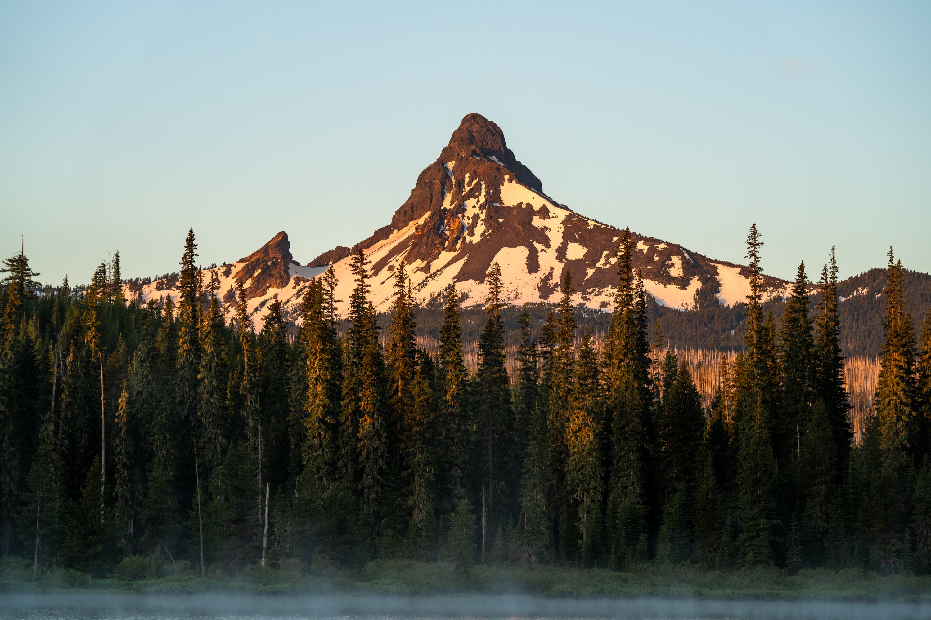 Snow-capped mountains behind a dense forest and misty lake HD Wallpaper