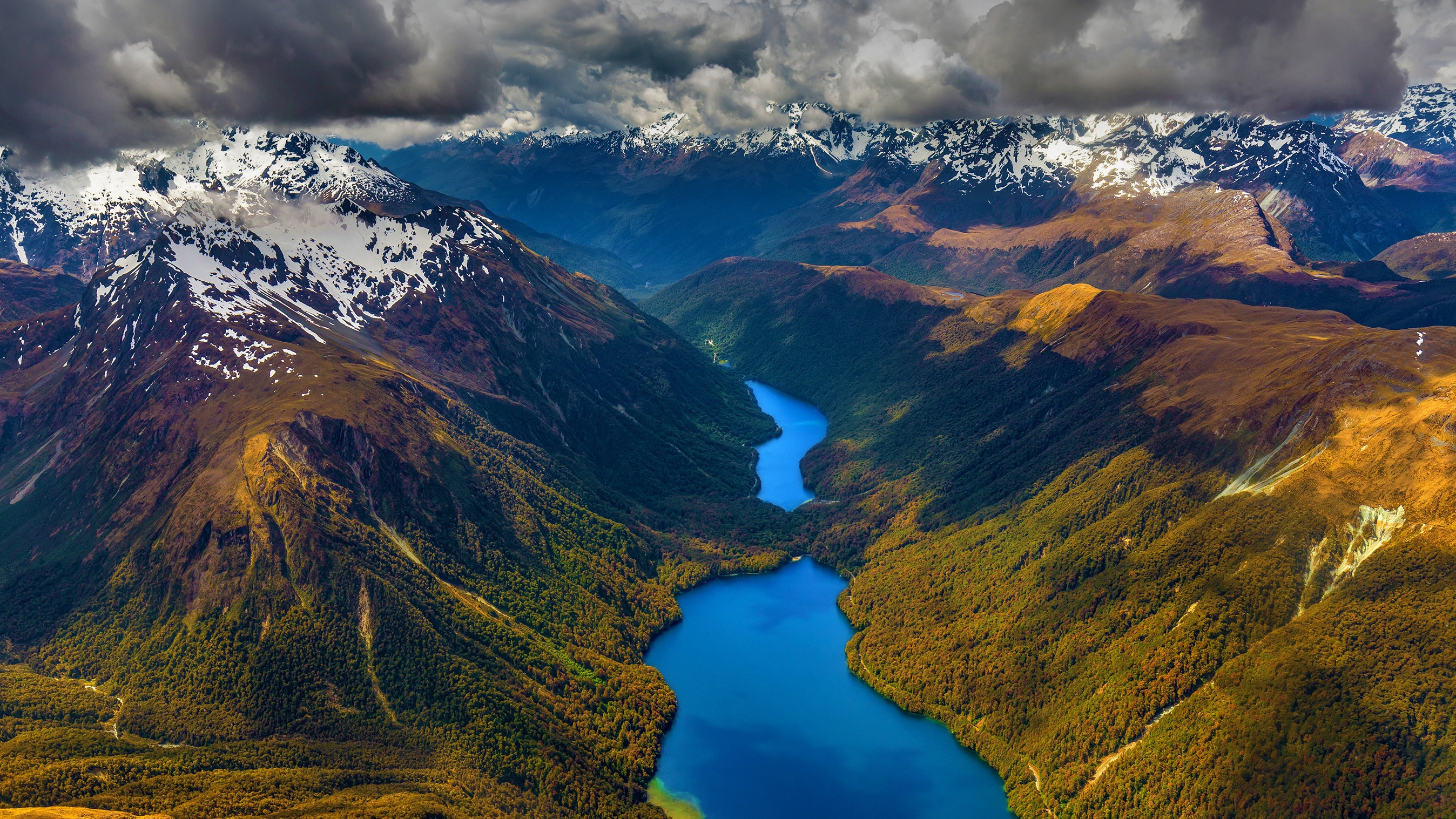 A winding blue river flows through a valley with snow-capped mountains HD Wallpaper