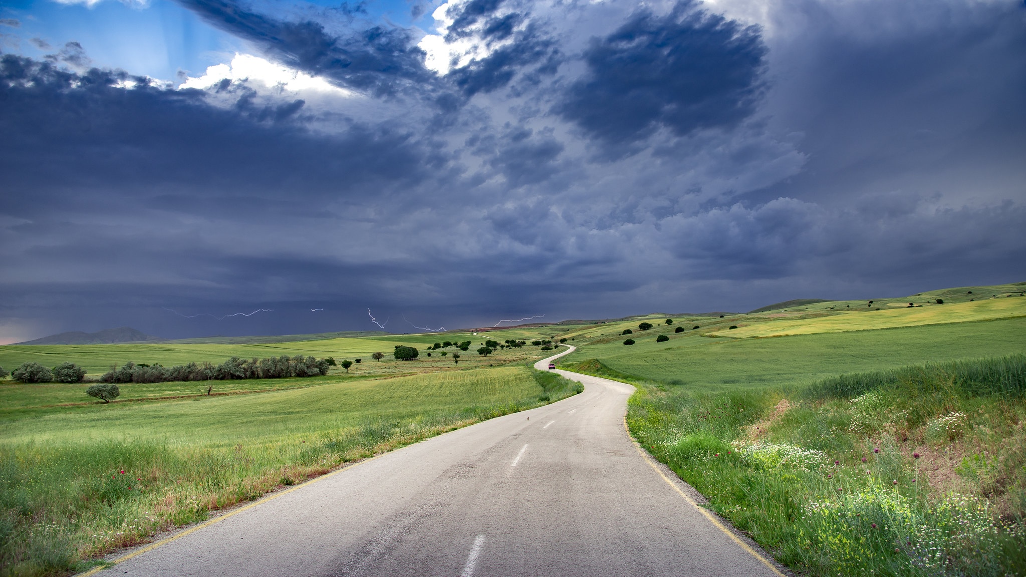 Road through green fields under stormy sky with lightning HD Wallpaper