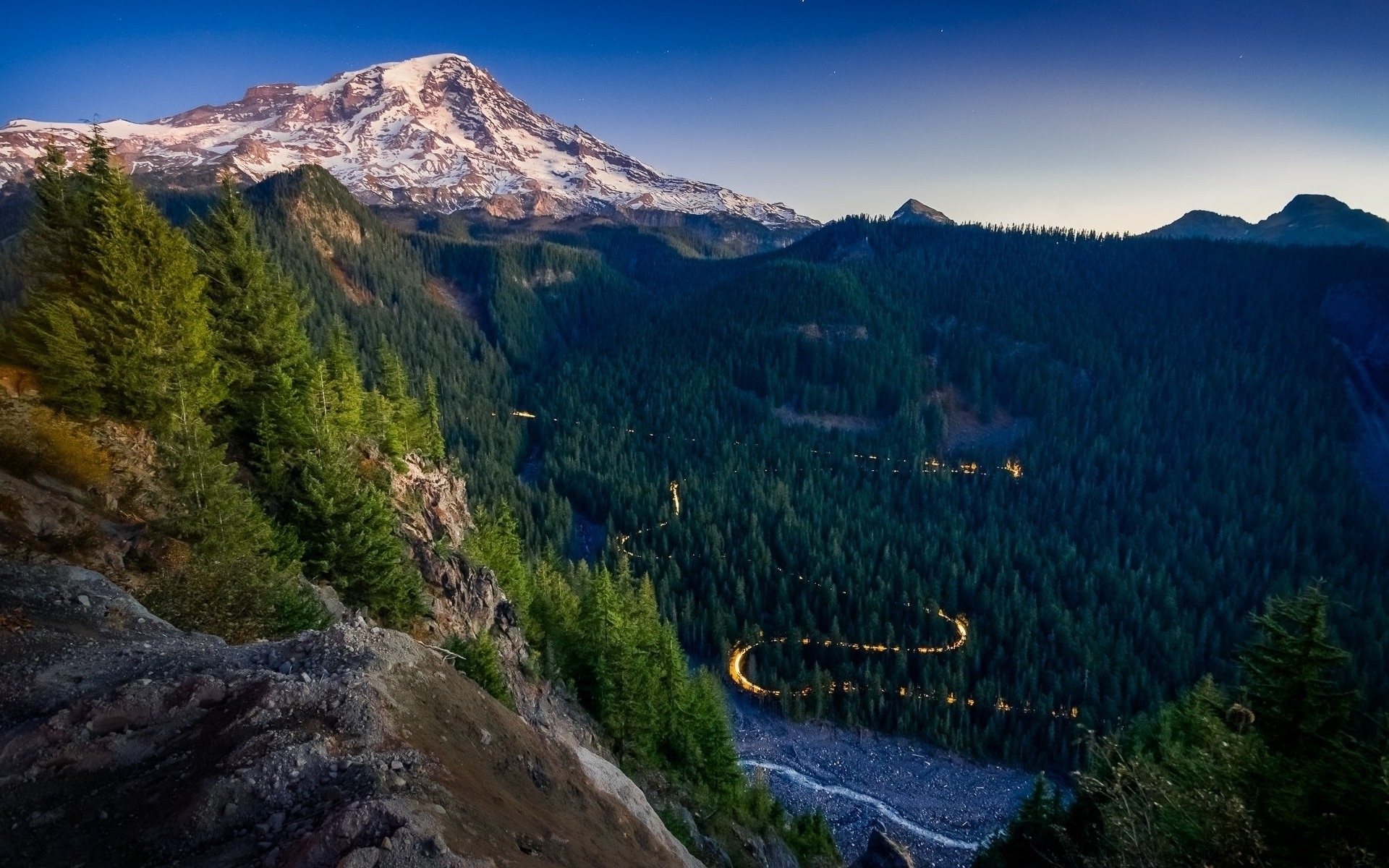 Snow-capped mountain overlooking a winding road through a forest HD Wallpaper