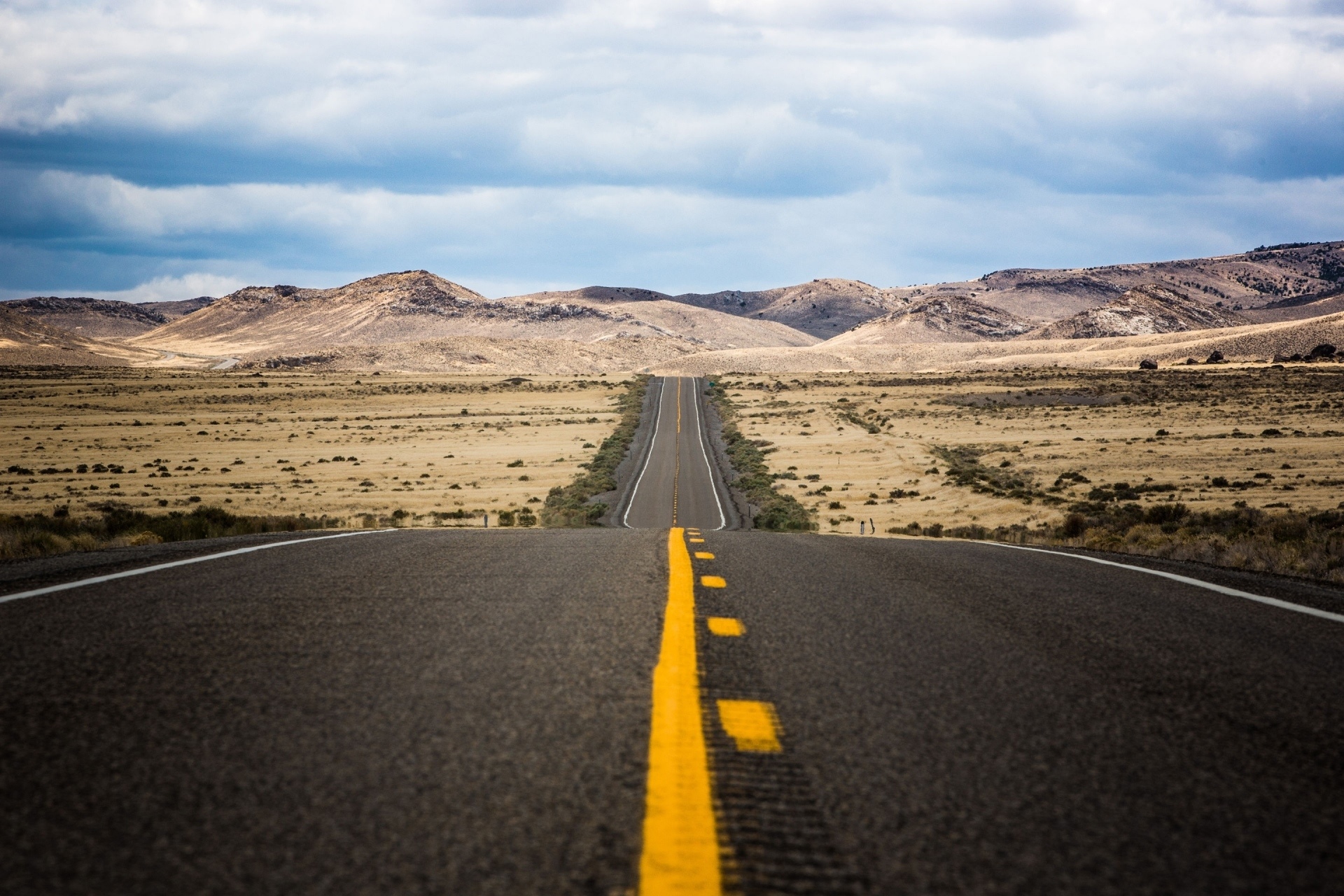 A long, empty desert road stretching towards mountains under a cloudy sky. HD Wallpaper