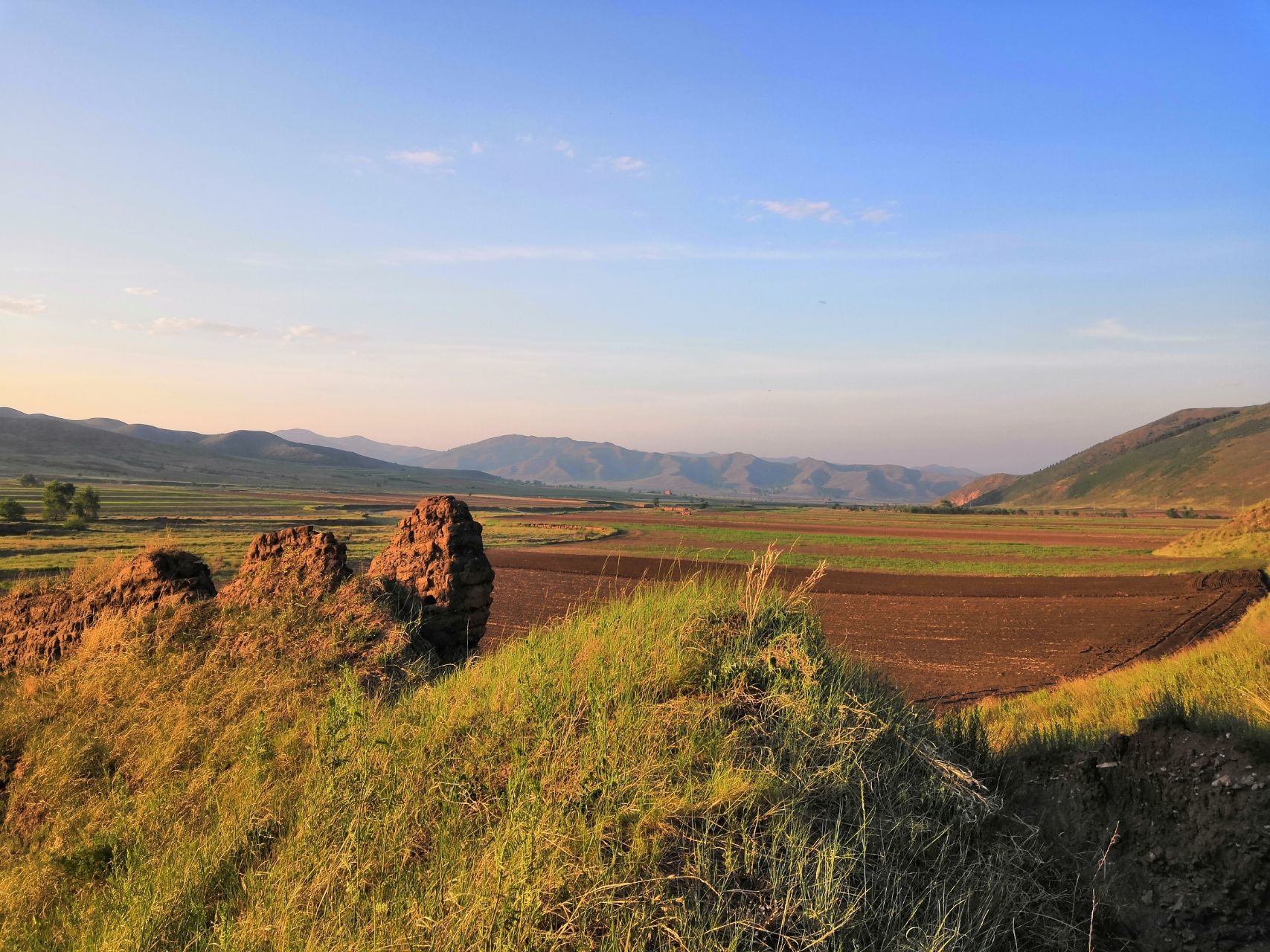 Landscape with fields, rocks, and mountains under a blue sky HD Wallpaper