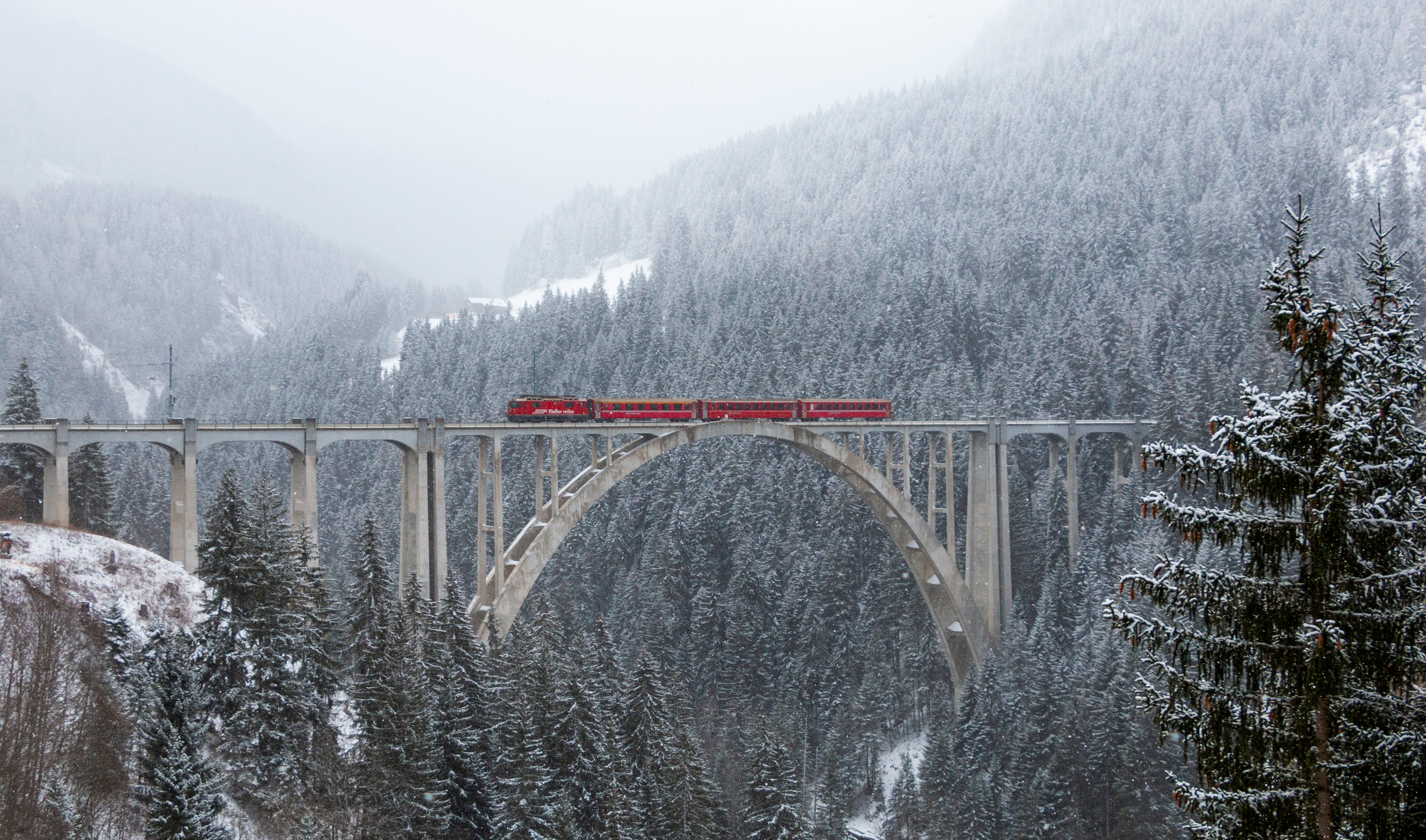 Red train crossing a snowy viaduct bridge in a winter forest HD Wallpaper